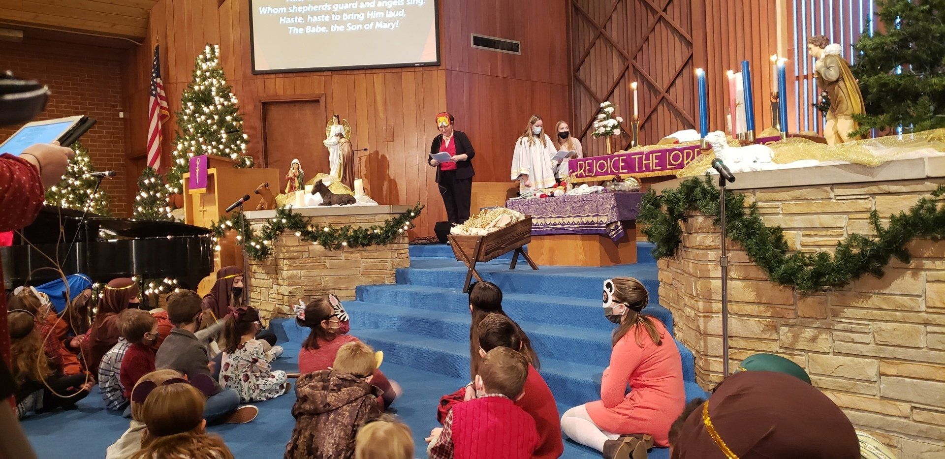 Children watch a Christmas play in a church, with a stage nativity scene and performers.