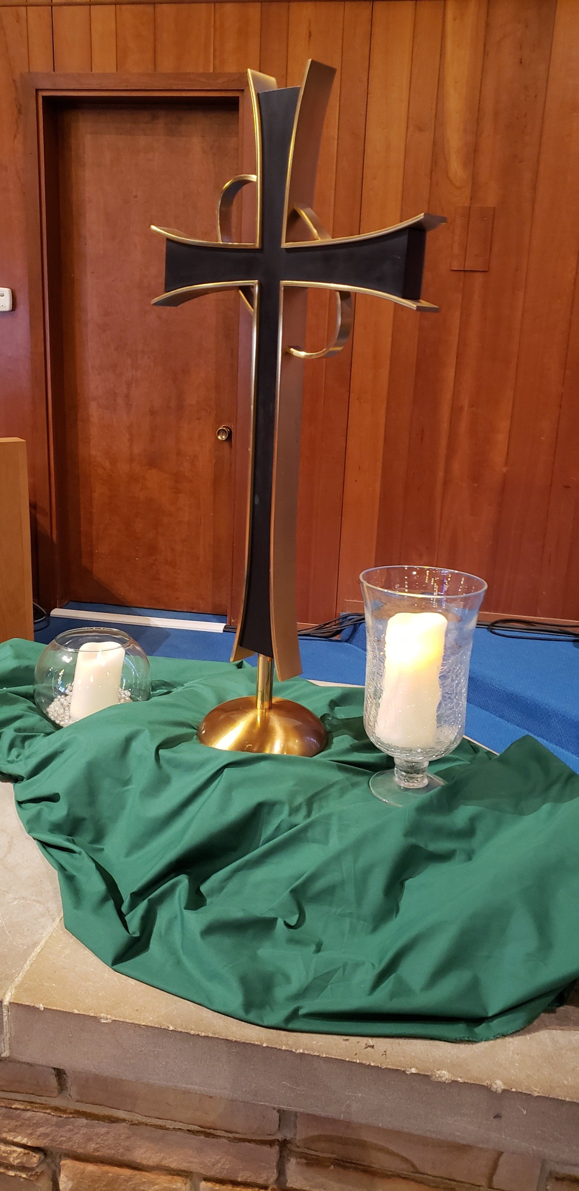 Black cross with gold trim on a table draped in green cloth, candles in the background.