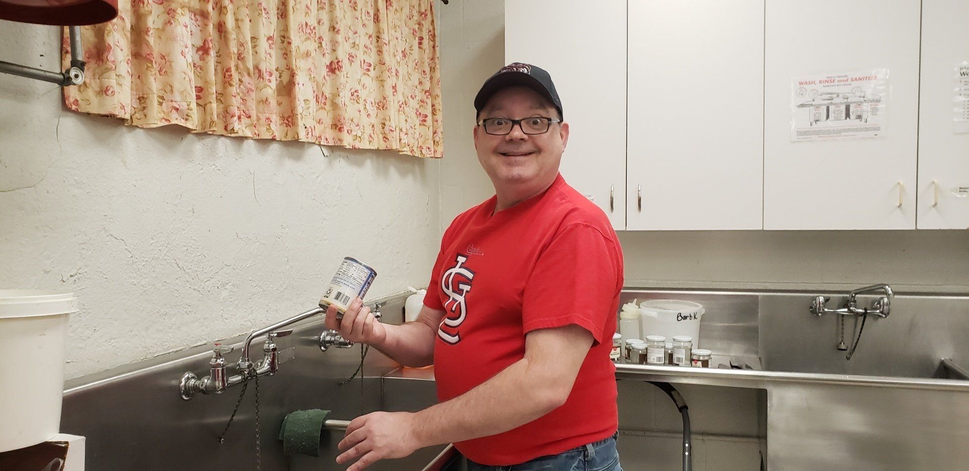 A man in a red shirt and cap smiles, holding a can. He stands at a stainless steel sink.