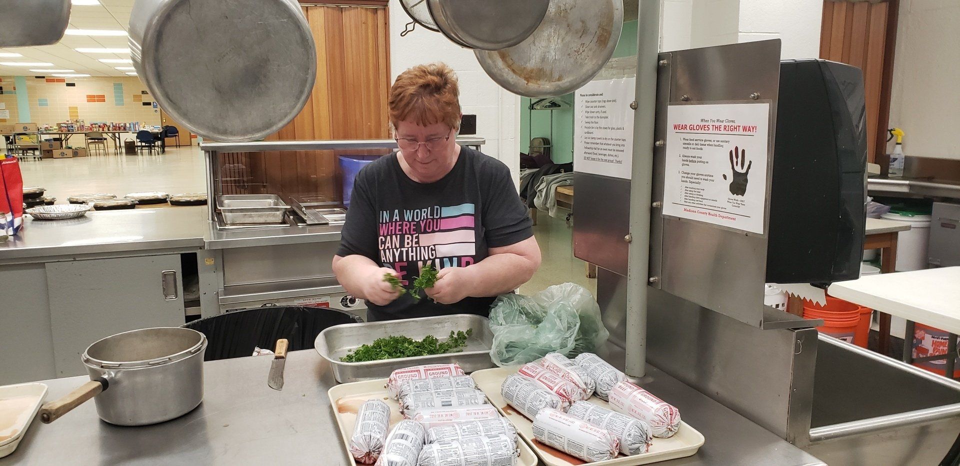 A person in a kitchen preparing greens. Wrapped items on trays, stainless steel surfaces, and hanging cookware.