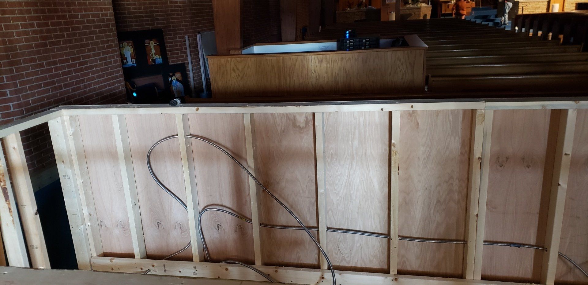 Interior construction of a wooden wall with exposed wiring, possibly a bar, with a brick wall visible.