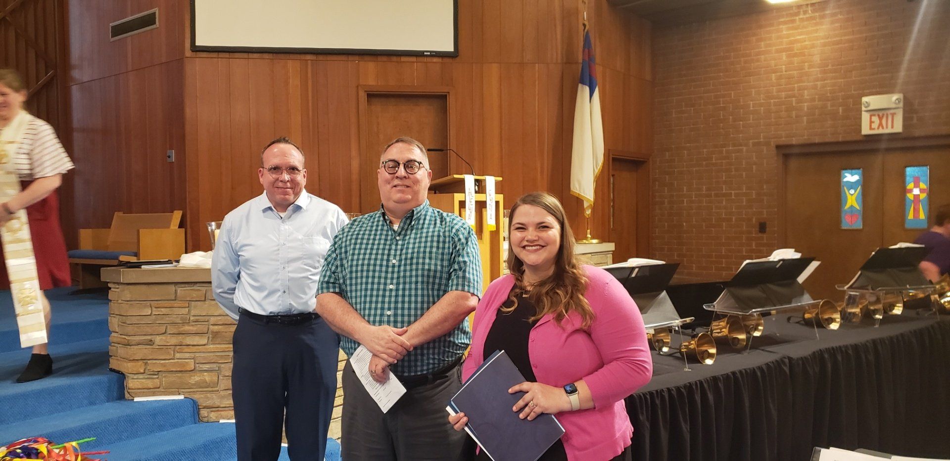Three people stand in a church. Woman in pink jacket holds a blue folder. Men in shirts, smiles. Bells on a table.