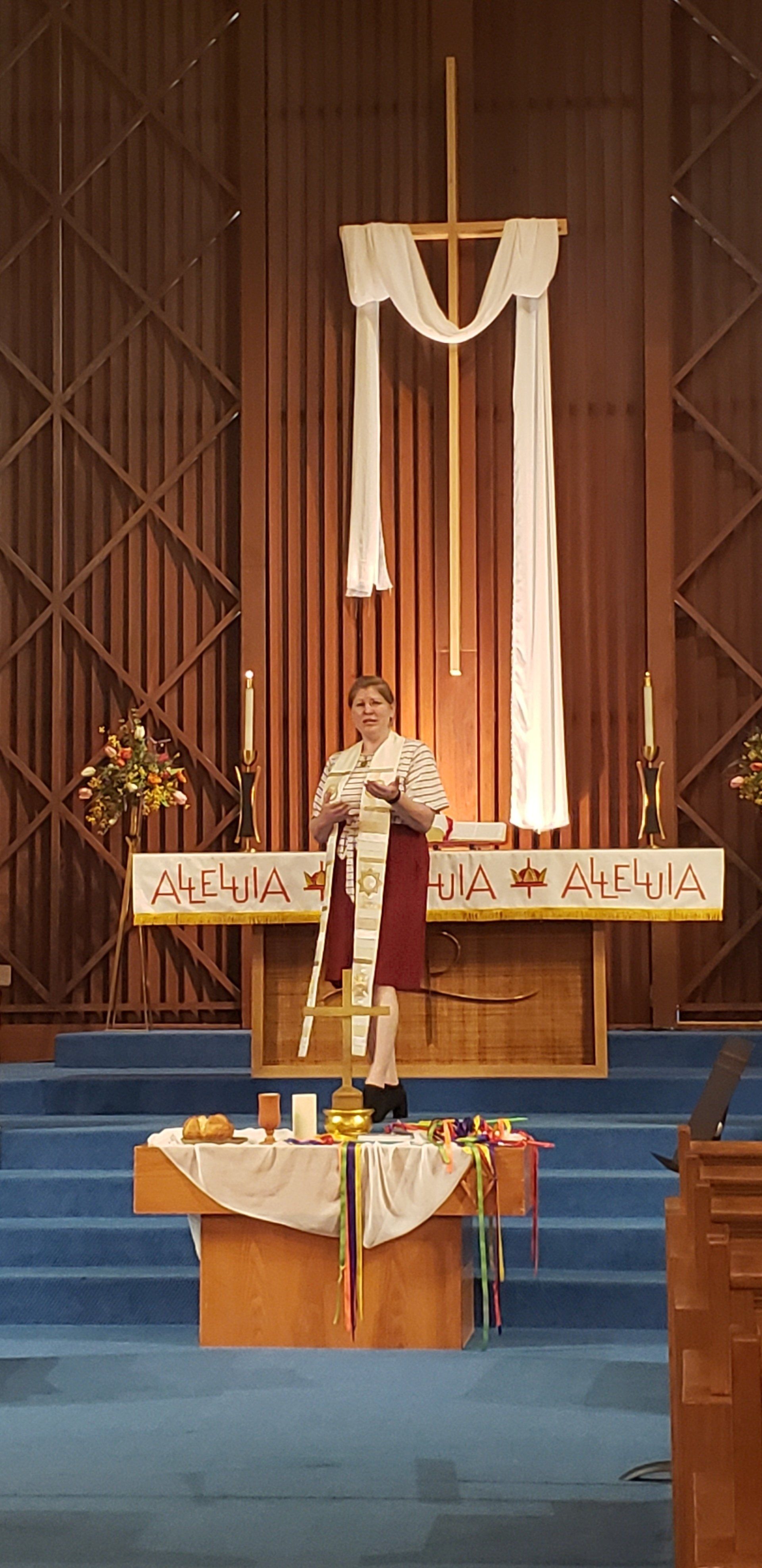 A person in religious attire stands at a church altar, speaking. Cross and draped cloth in the background.