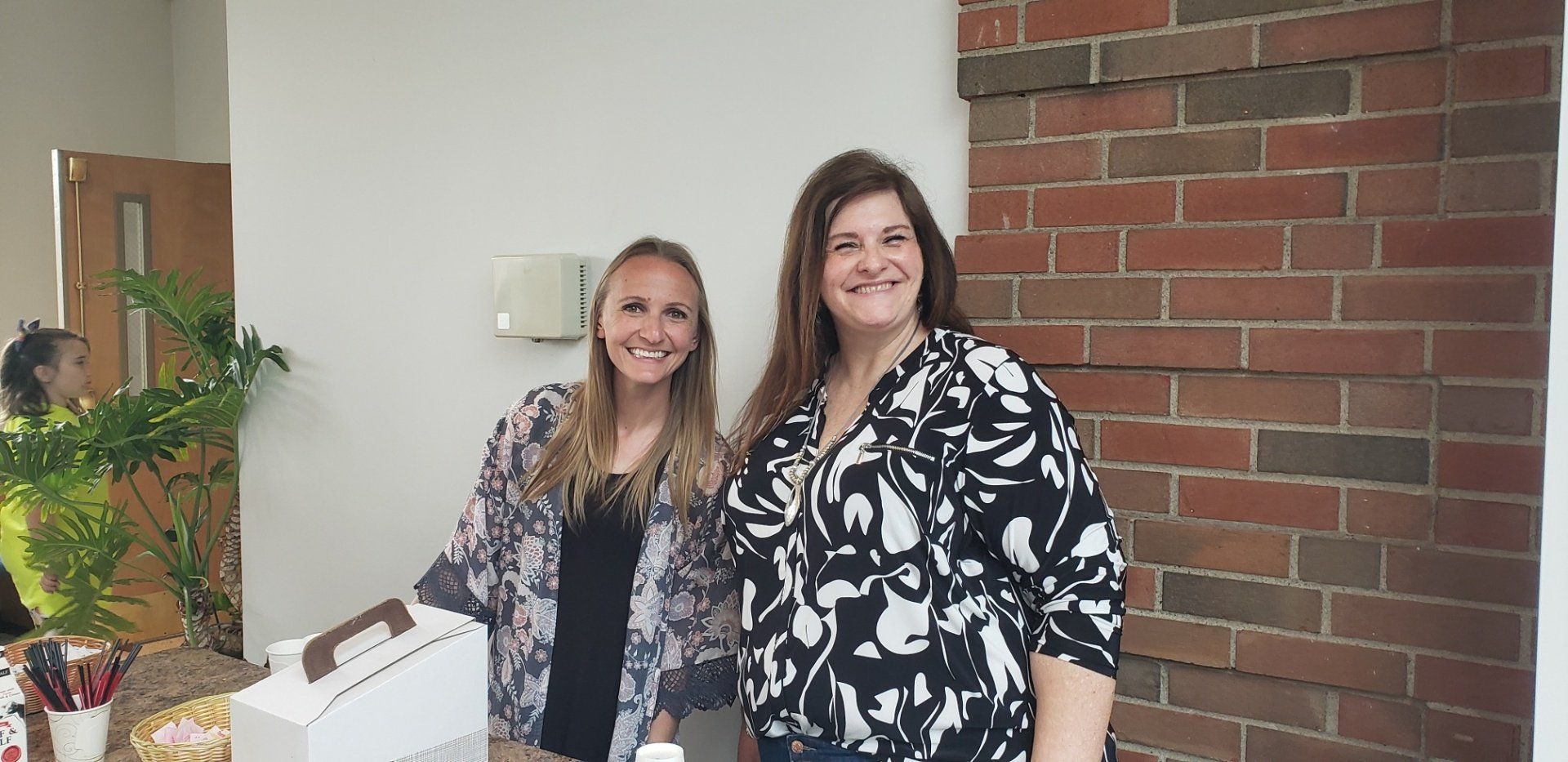 Two smiling women stand near a brick wall; one in a floral top, the other in a patterned jacket.