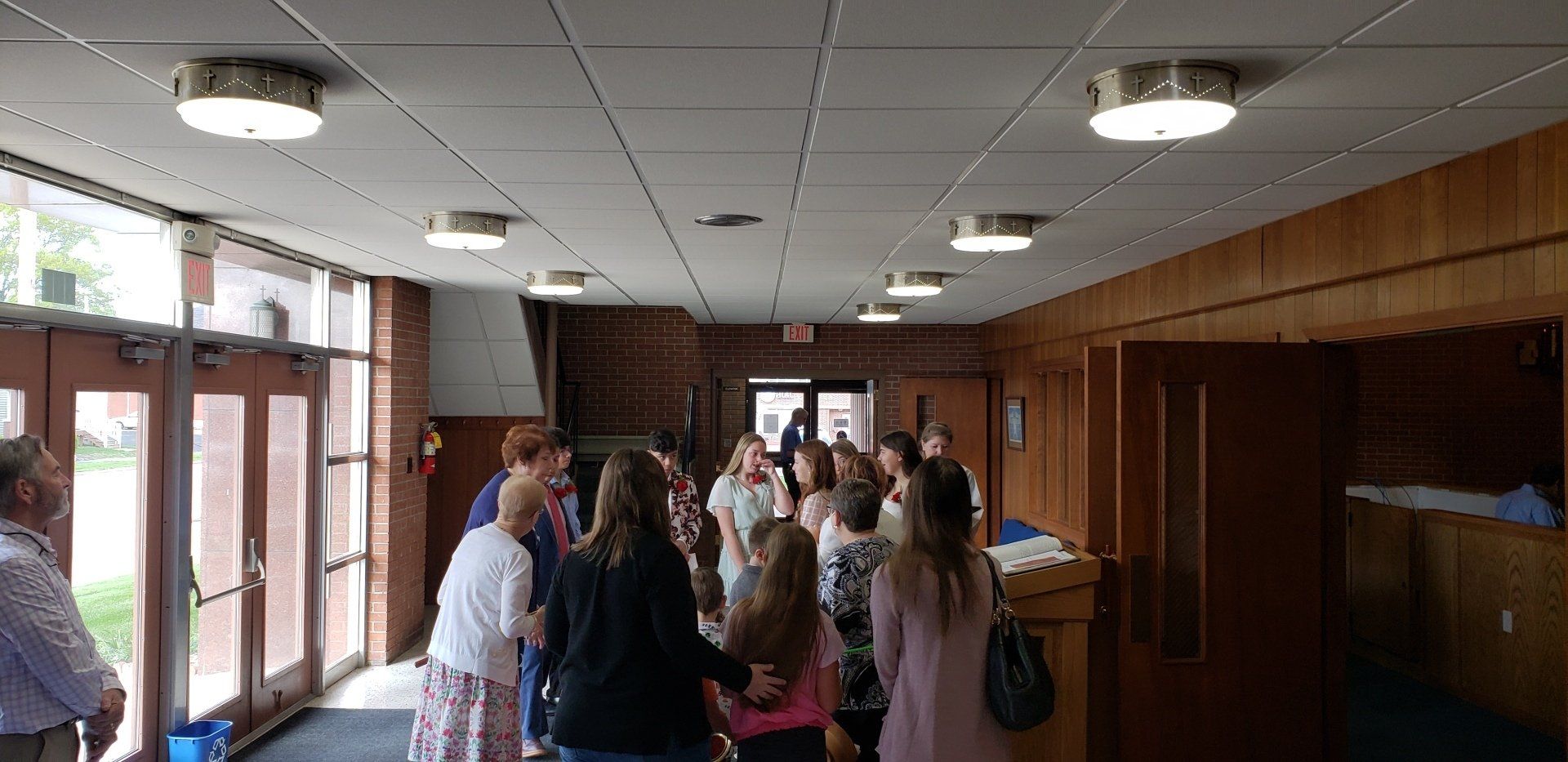 People gather inside a hallway, near glass doors. Wood paneling, ceiling lights.