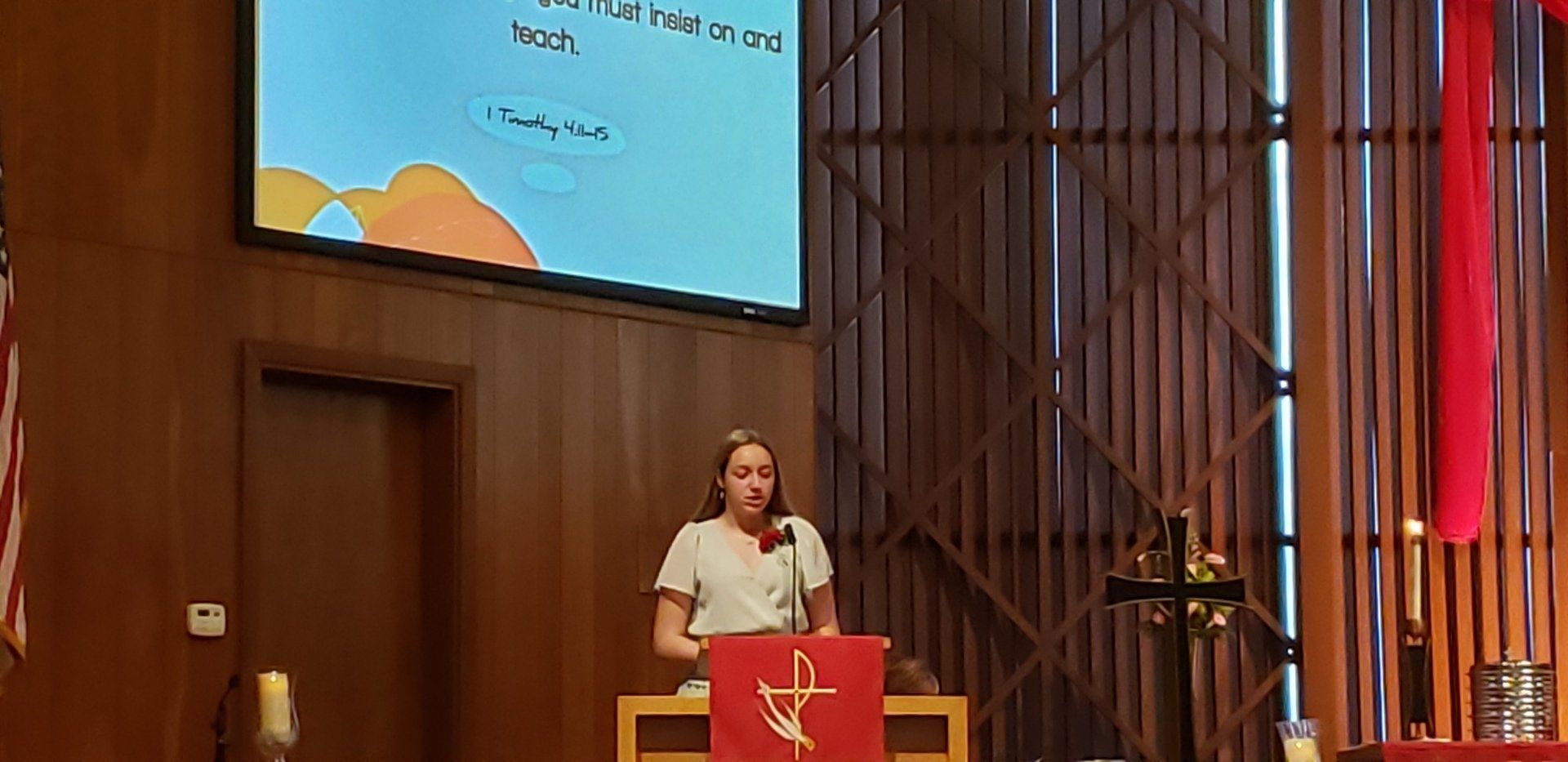 Woman speaking at a lectern in a church. Screen shows a title. Red fabric drapes nearby.