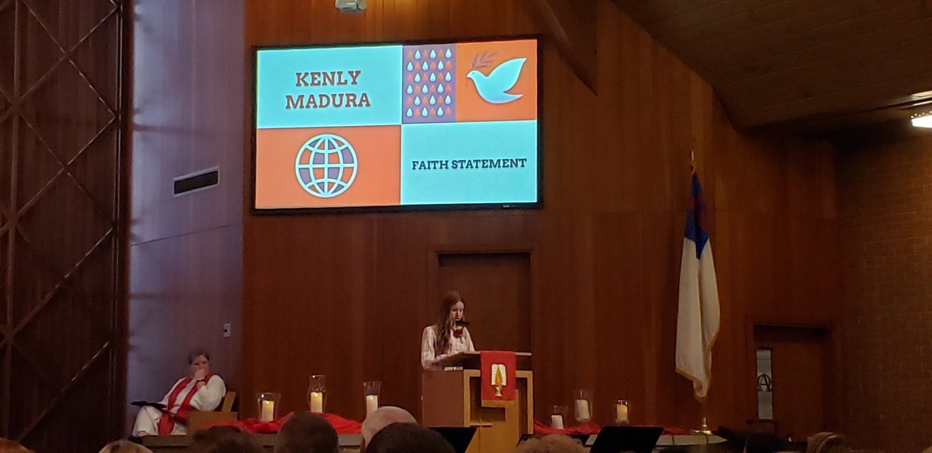Woman speaking at a podium in a church. A screen displays words and images, including a dove. A Christian flag is present.