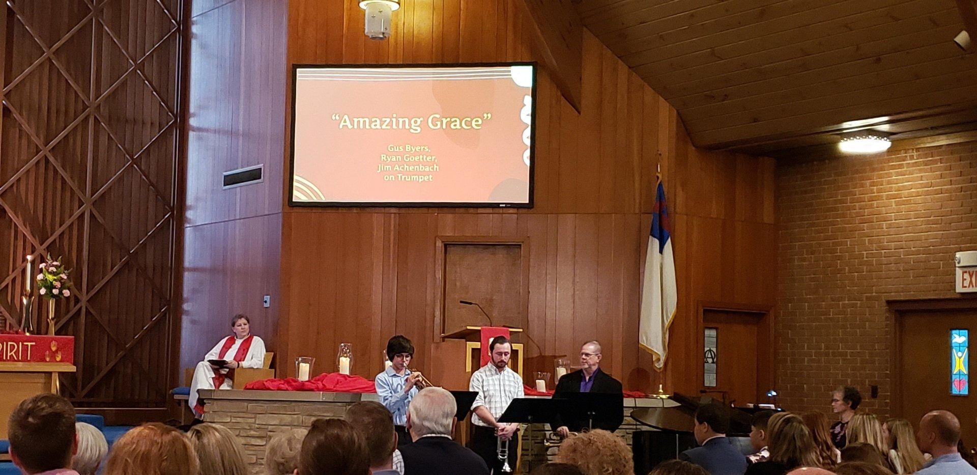 A church service with a man speaking and two children, people seated. Interior with wooden paneling.