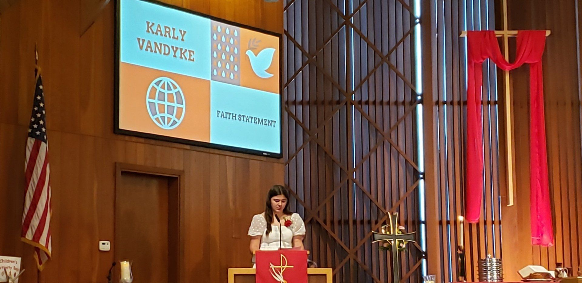 A woman speaking at a podium in a church with a presentation screen and American flag.
