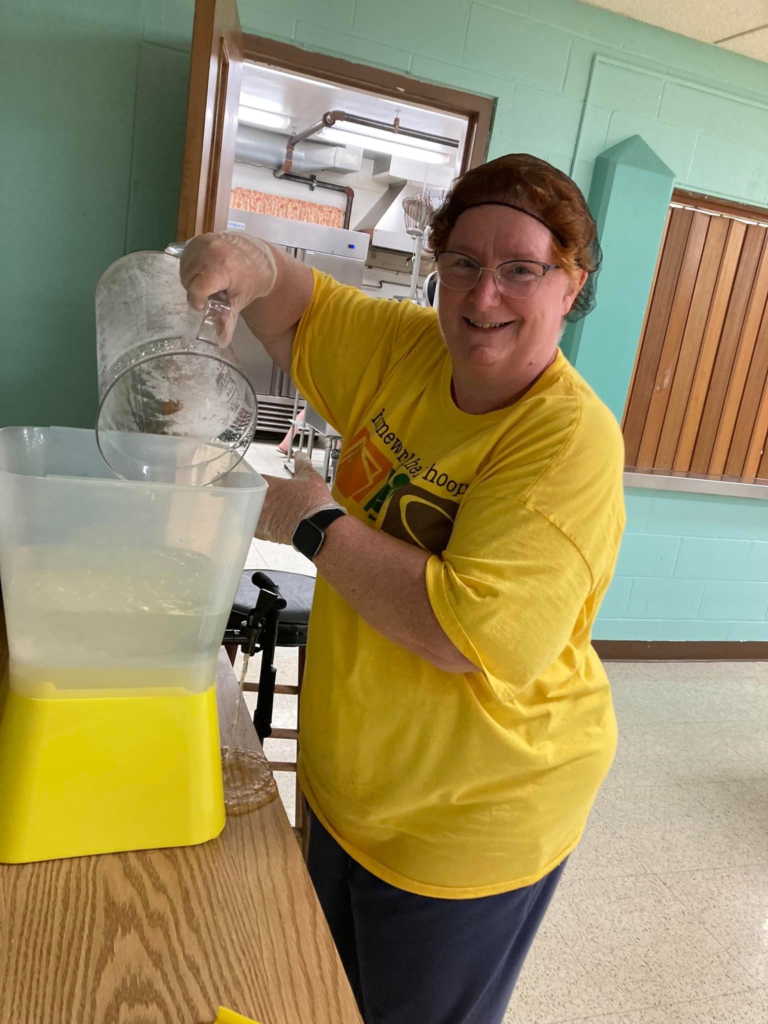 Woman in yellow shirt pouring liquid into a yellow dispenser; indoor, smiling.