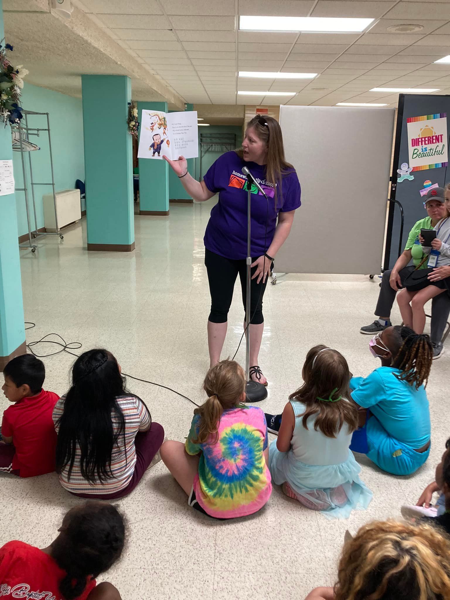 A woman reads to a group of children sitting on the floor in a hallway; she holds a book.