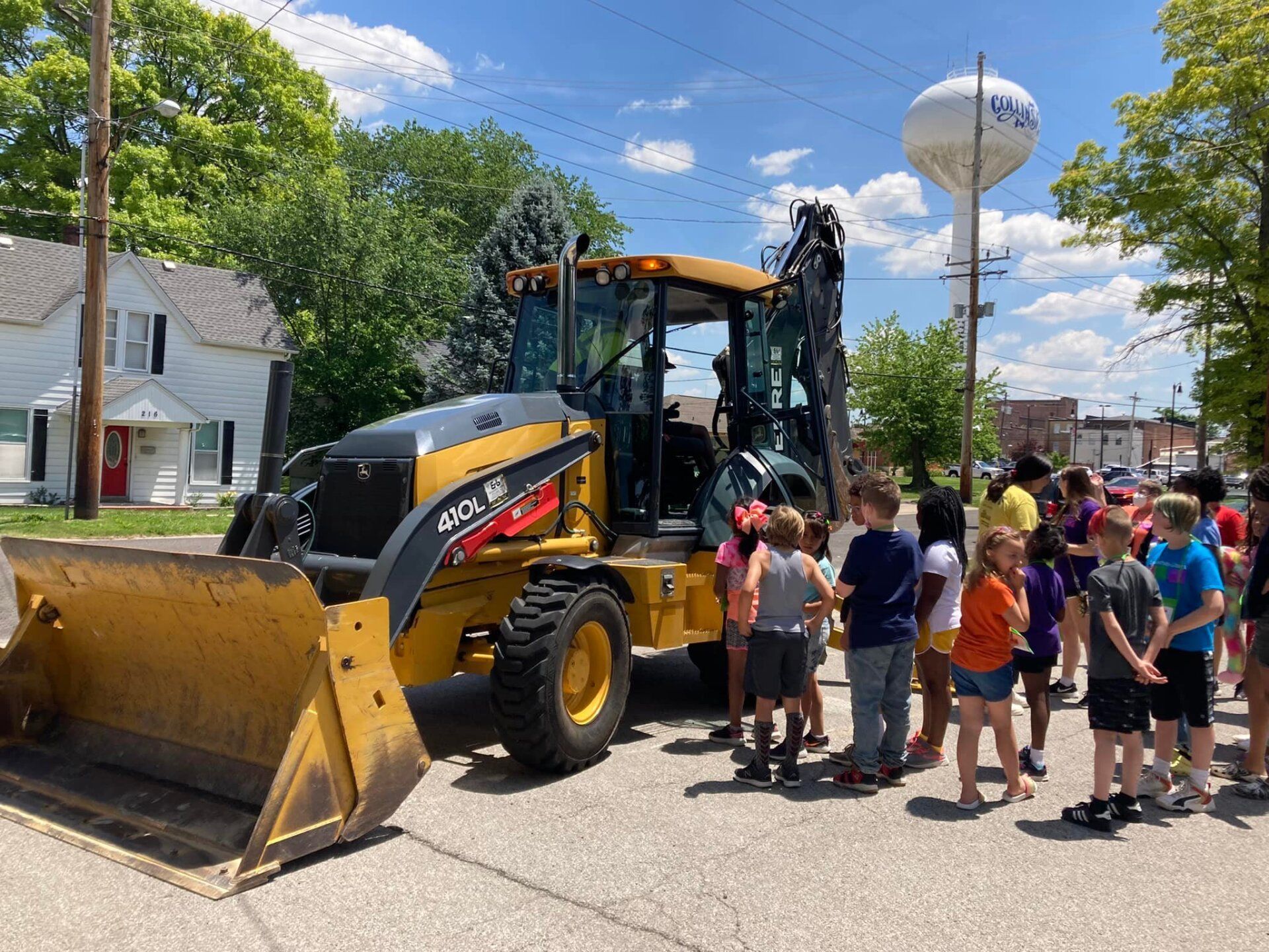 Children gather around a yellow construction vehicle on a street, under a blue sky.
