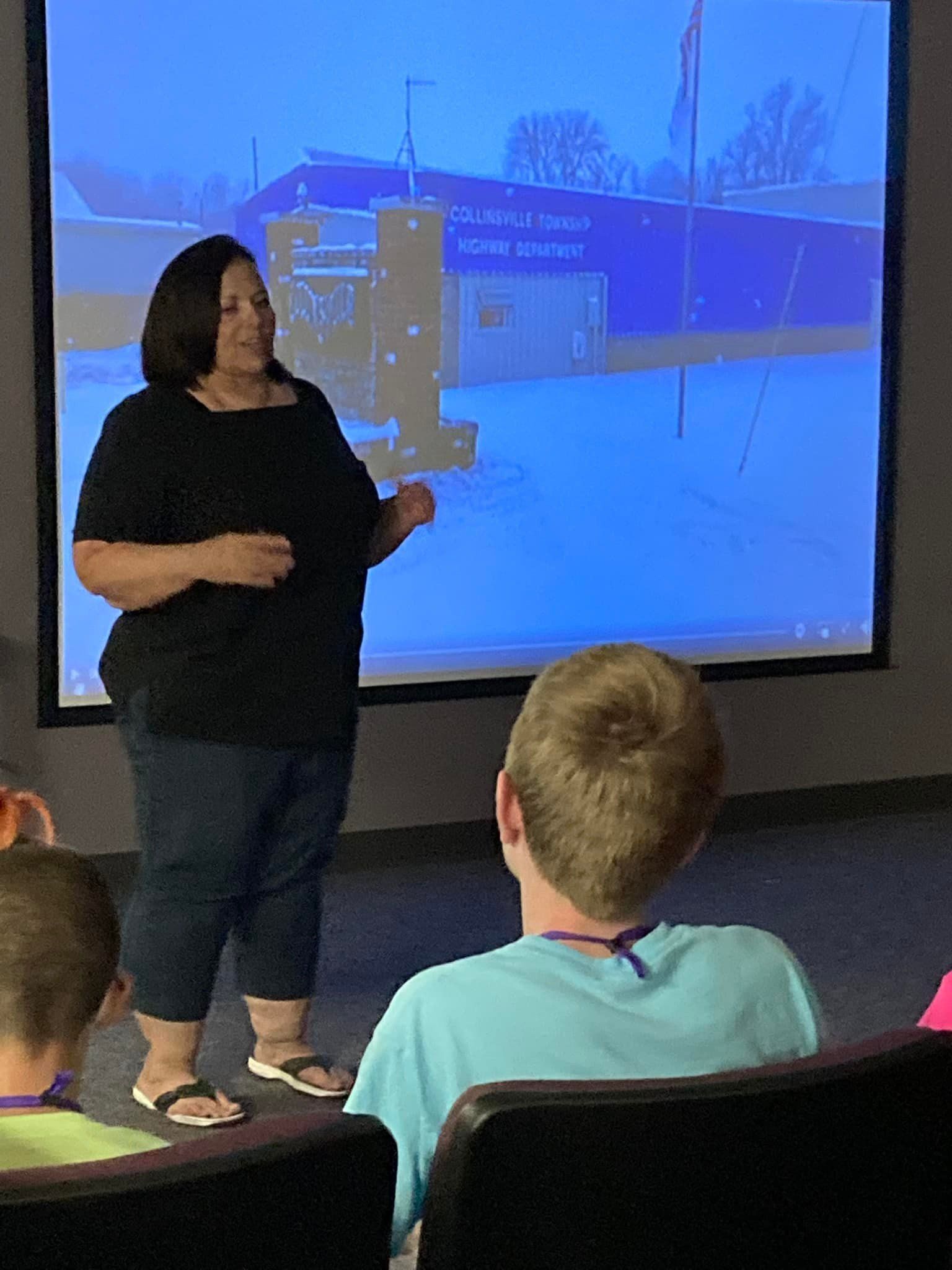Woman presenting in front of a screen. She gestures with her right hand. A young person sits in the audience.