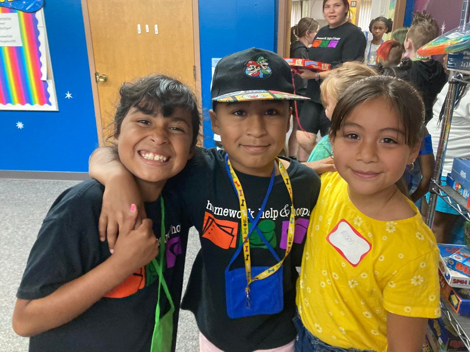 Three smiling children with arms around each other inside a building, brightly colored room.