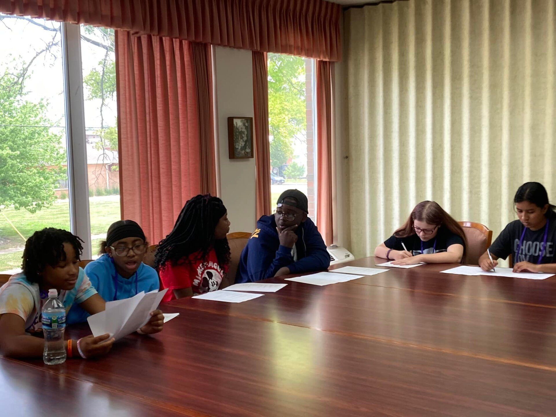 A group of teens sit around a table reading papers in a well-lit room with curtains and a window.