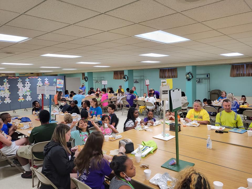 Children and adults seated at tables, eating lunch in a large room with fluorescent lights.