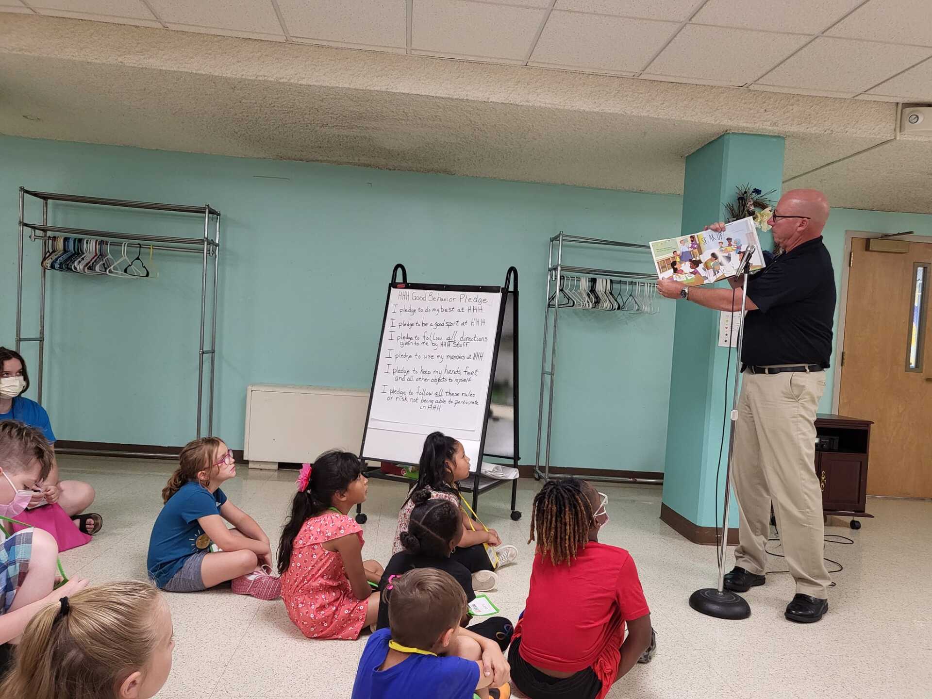 Man reading a book to a group of children sitting on a floor in a room with a teal wall.