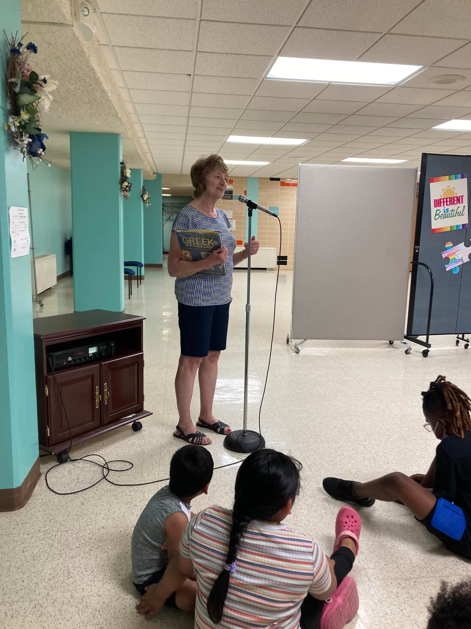 Woman reading to children, indoors. She holds a book, speaks into a microphone. Children sit on the floor.