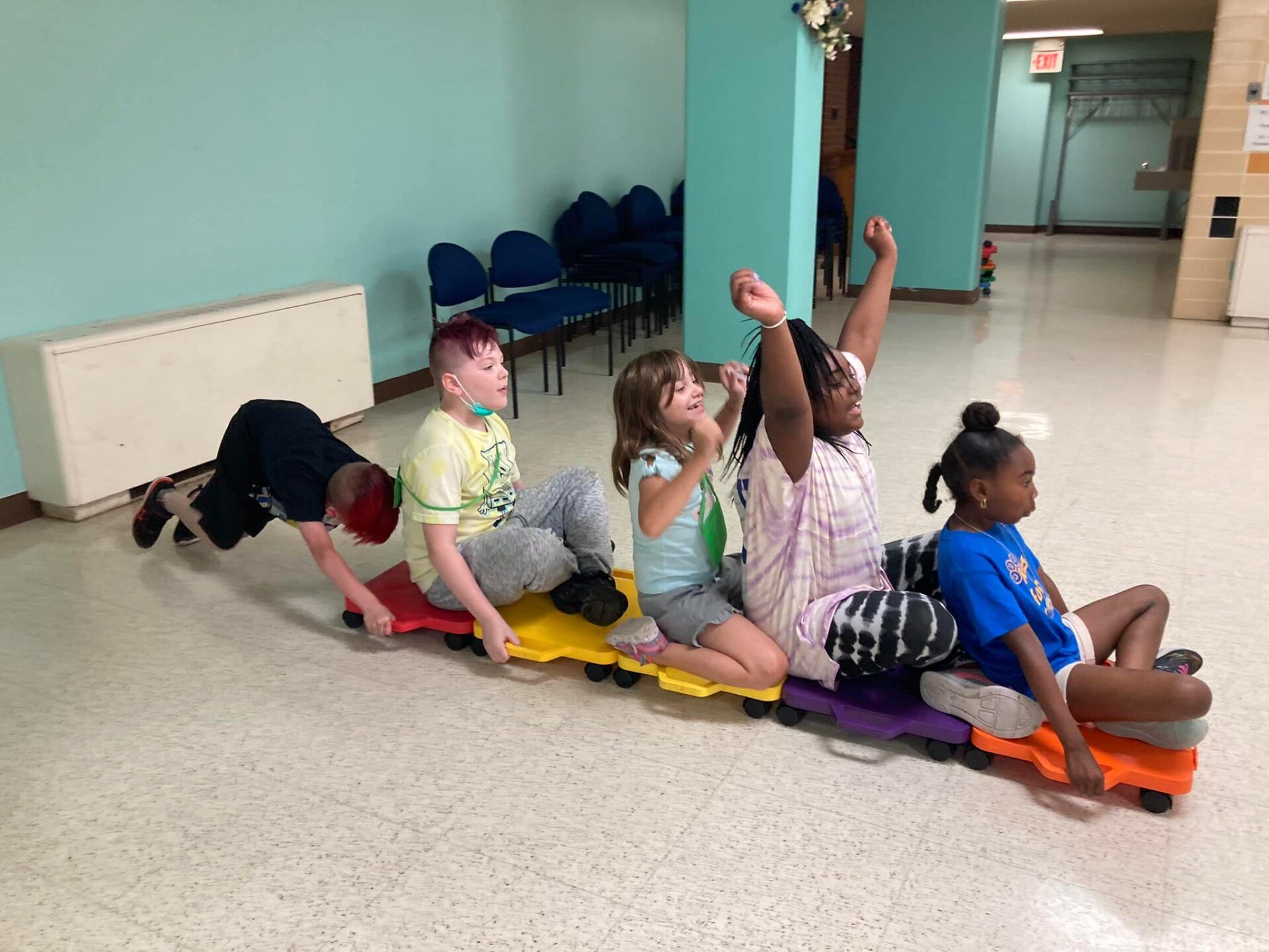 Children riding a colorful train of scooters indoors, with one child pulling.