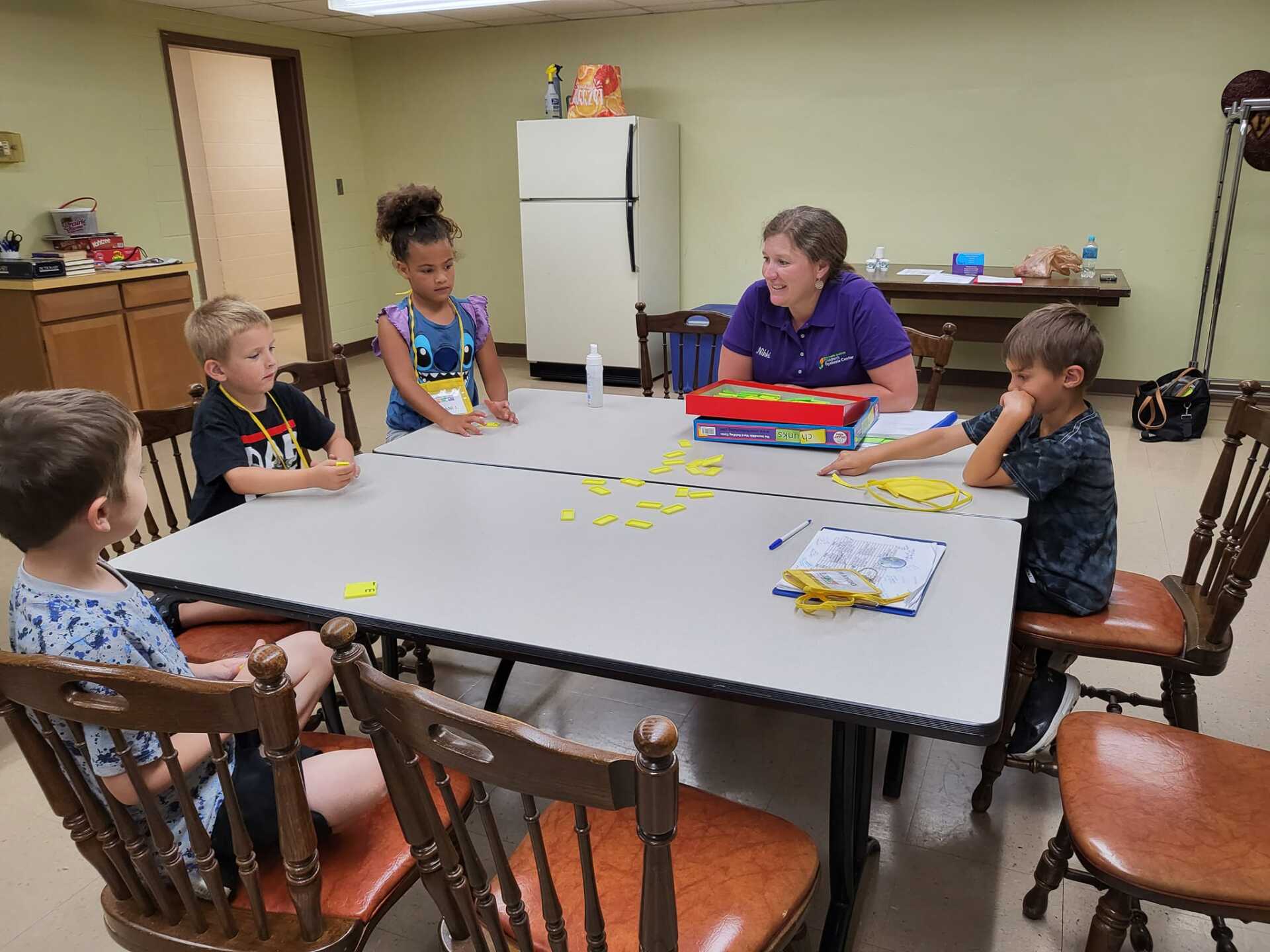 Children and adult playing a game at a table.  A woman in purple looks at the children. A table with yellow markers.