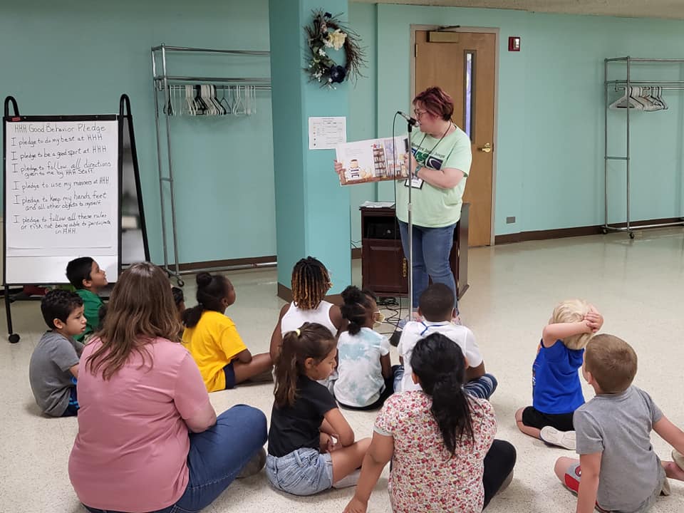 A teacher reads to children sitting on the floor. A whiteboard with writing stands nearby.