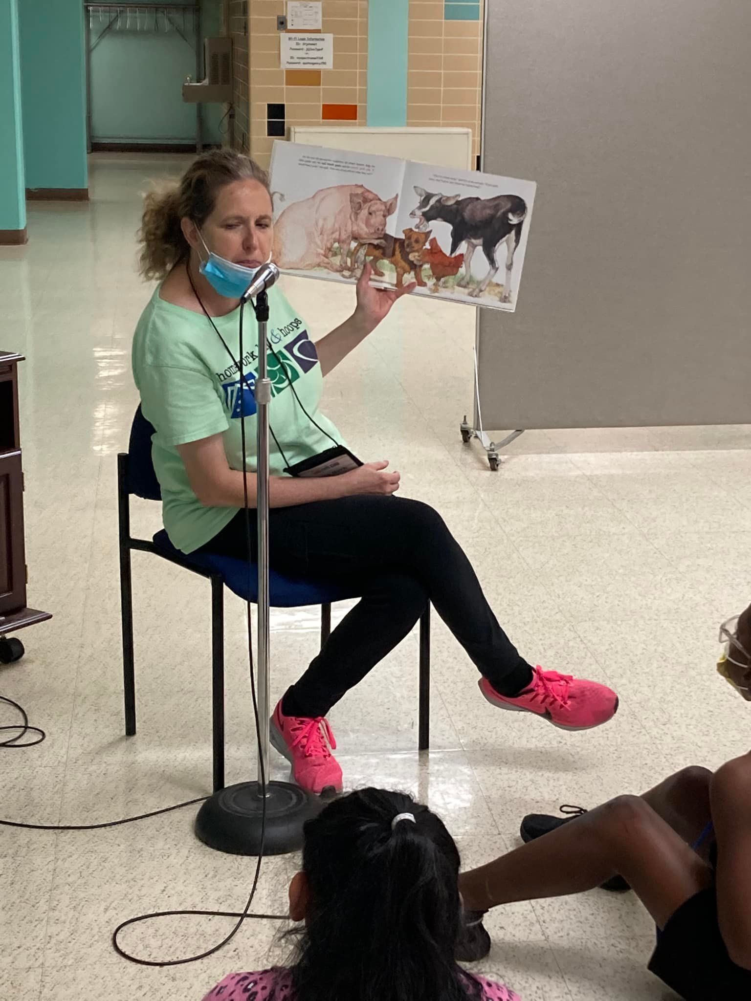 Woman reads aloud from a picture book to children. She wears a mask and pink shoes, seated.