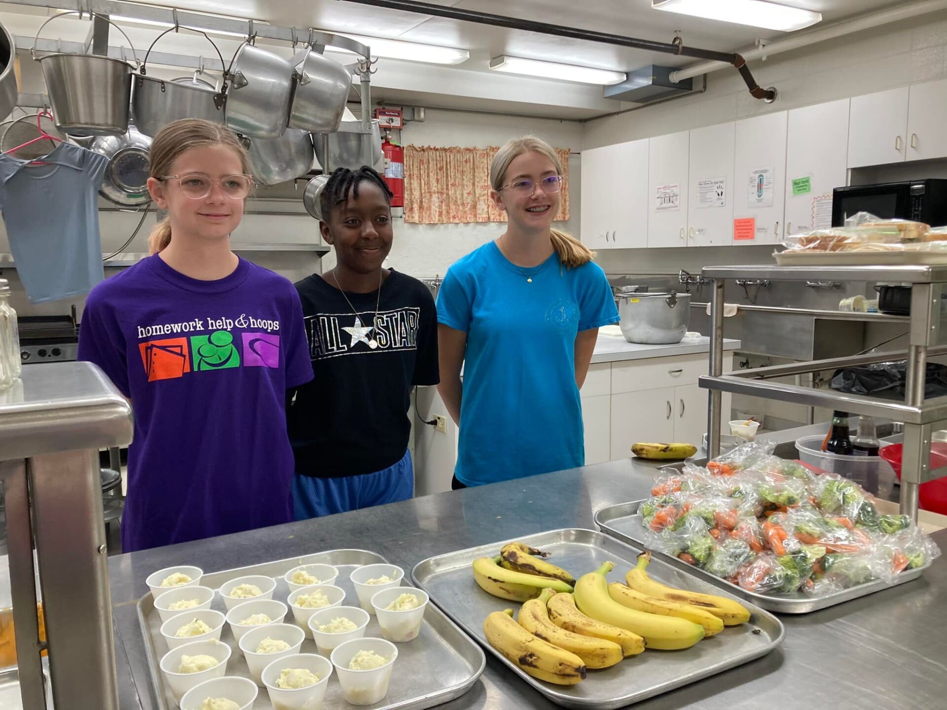 Three girls stand in a kitchen near trays of bananas, yogurt cups, and wrapped vegetables.