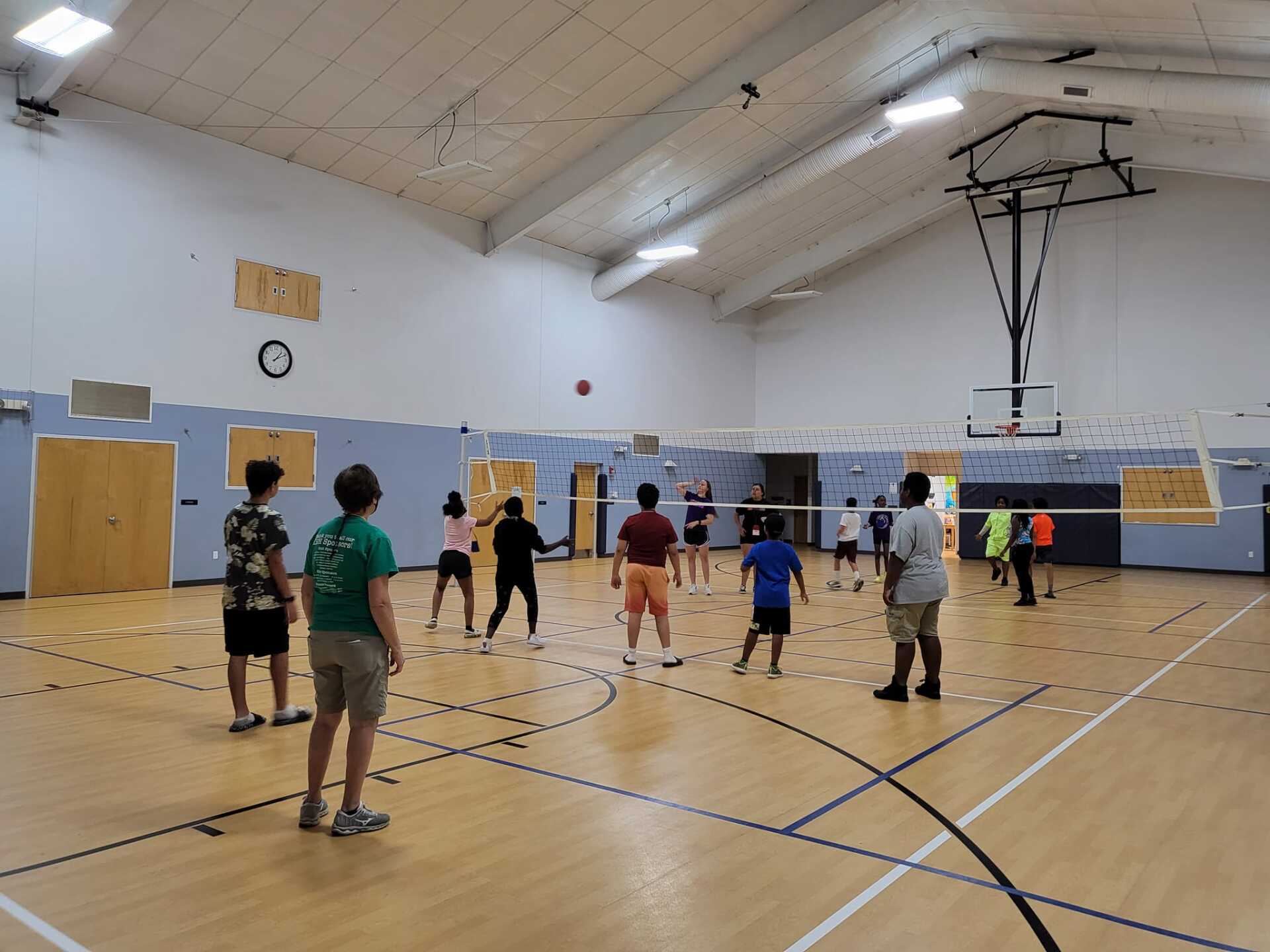 People playing volleyball in a gymnasium. A ball is mid-air above the net.
