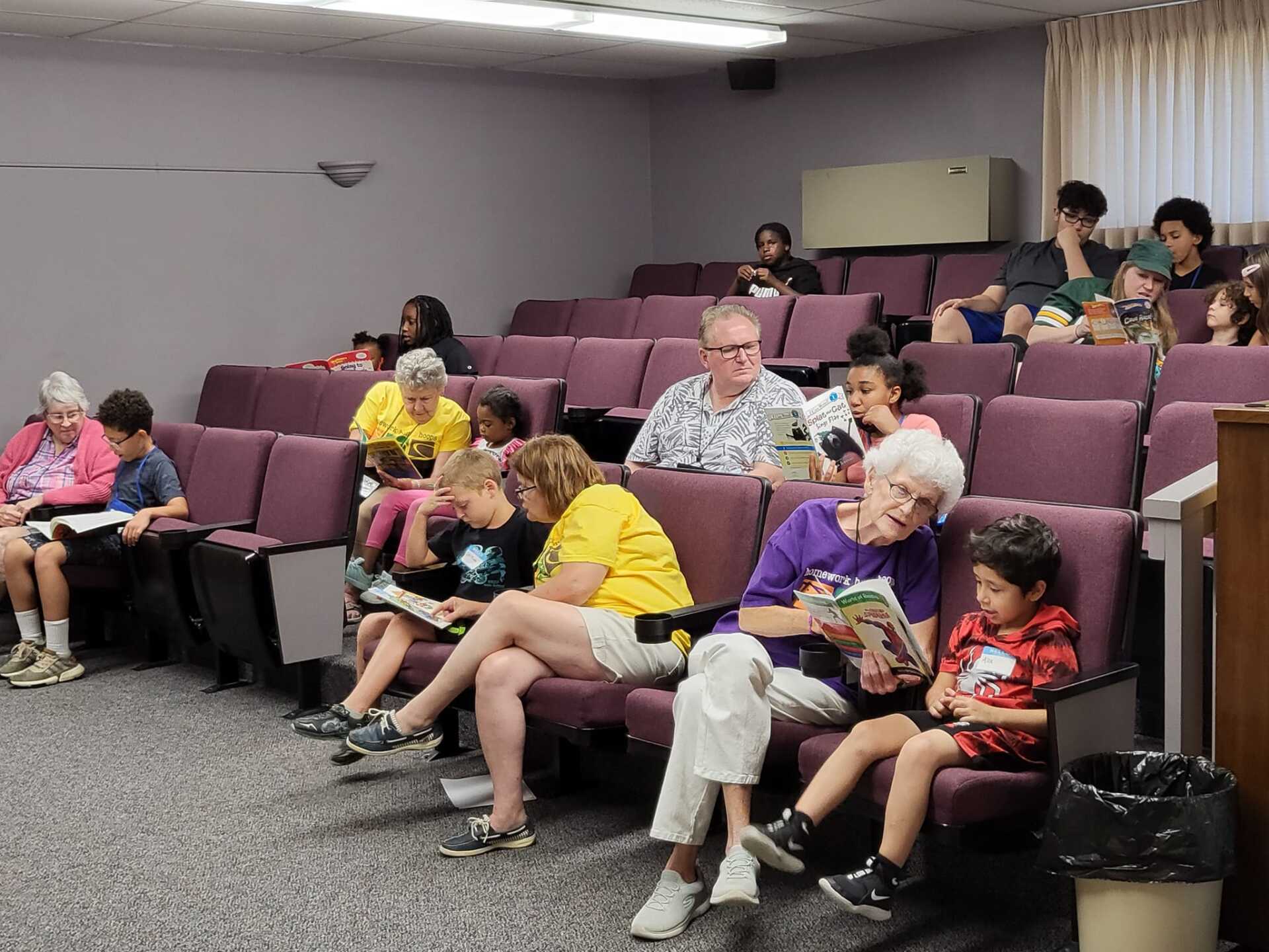 People of various ages reading books in a theater-style room with purple seats.