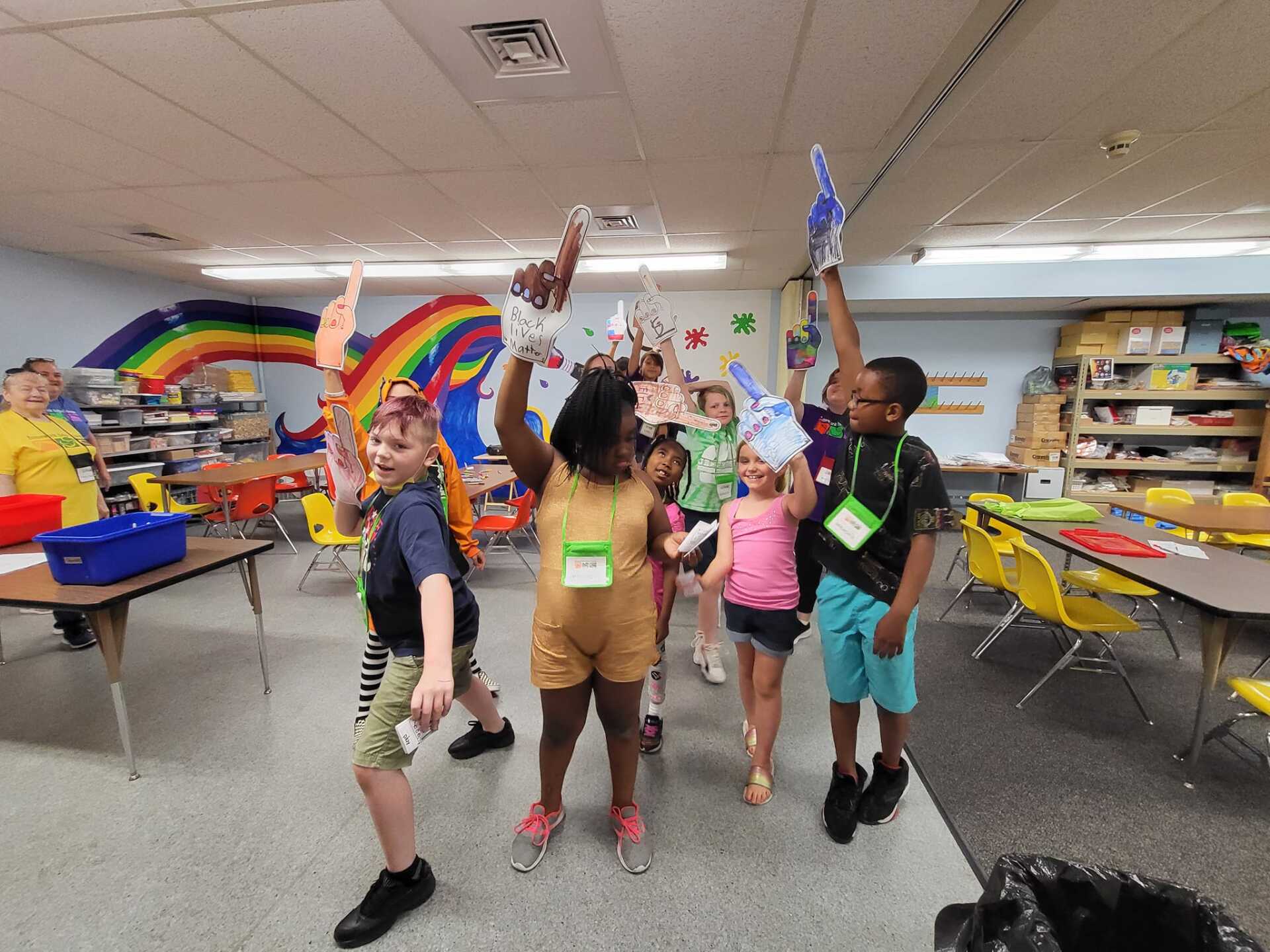 Children in a classroom celebrate, arms raised, holding artwork, with a rainbow mural in the background.