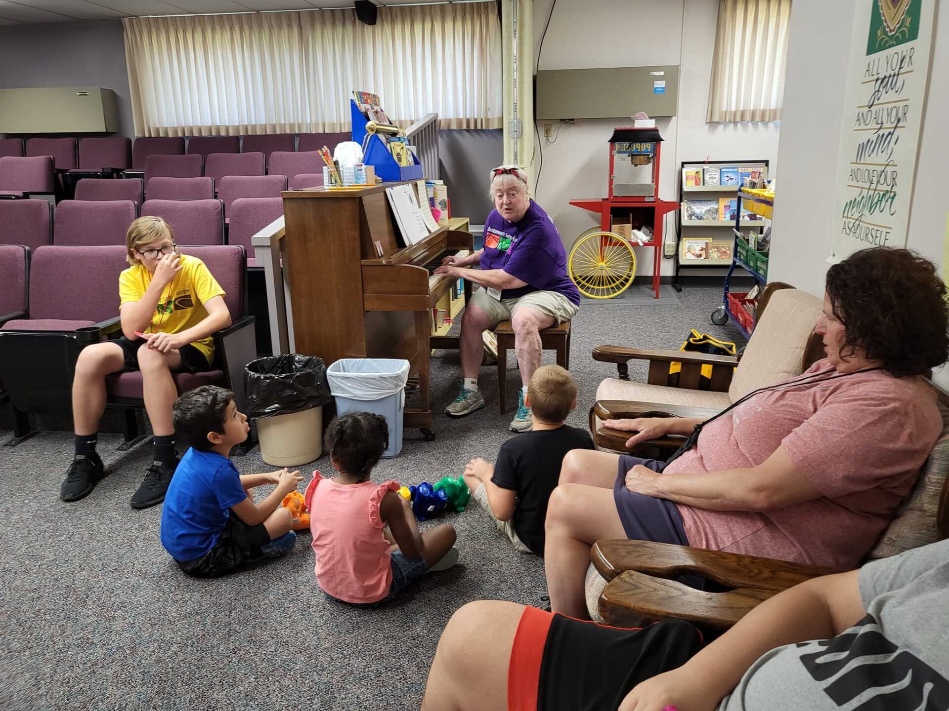 Children and adults in a room with a piano; one person plays, others watch.