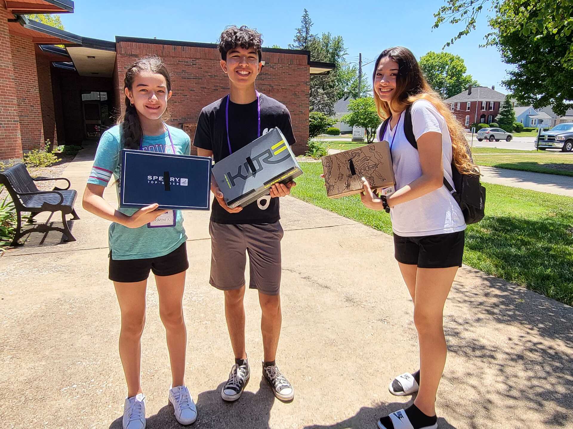 Three smiling teens holding DIY laptop covers outdoors. Brick building and sunny day.