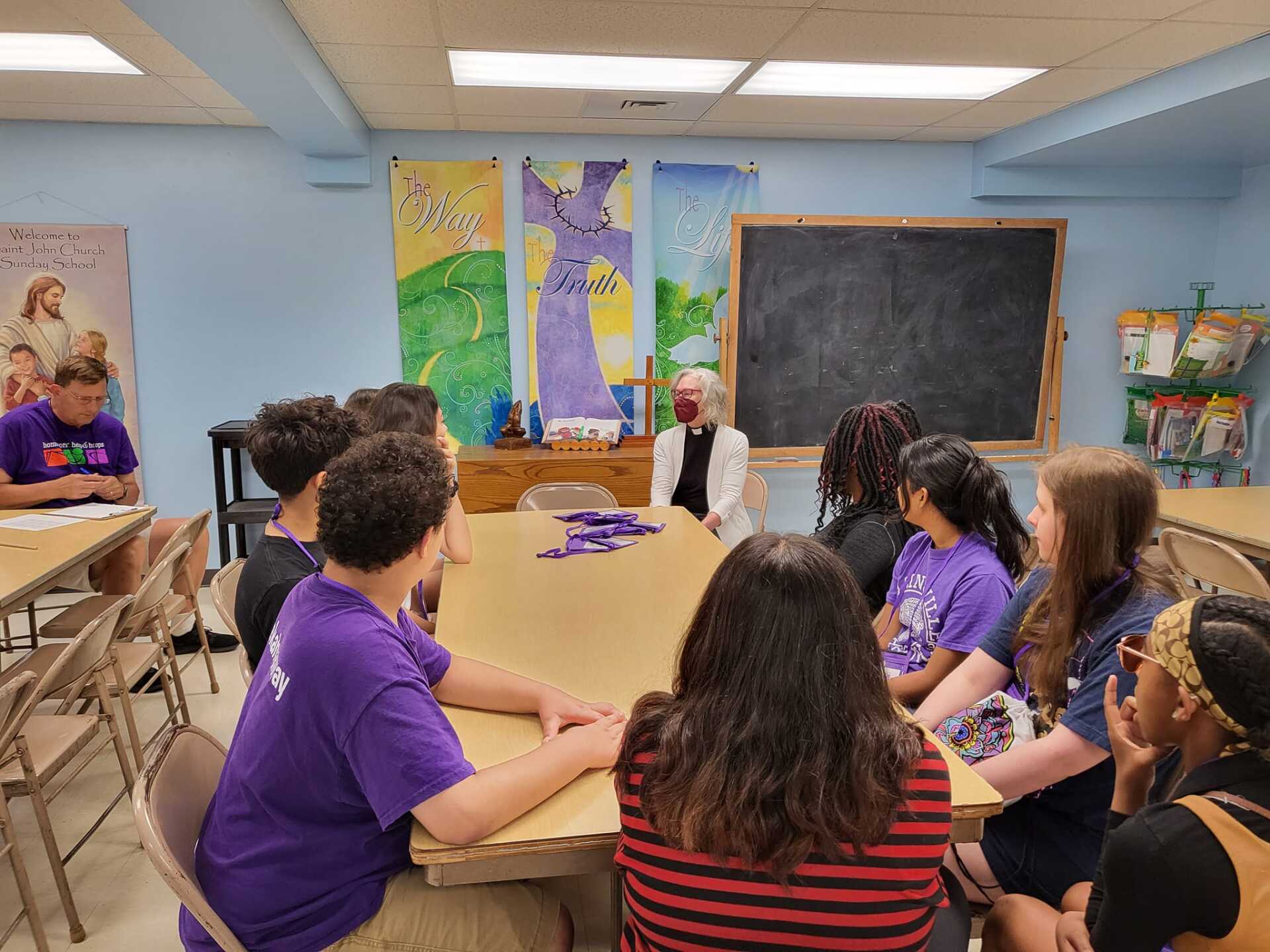 A group of teens in purple shirts sit around a table, talking with an older woman in a room with art and a chalkboard.