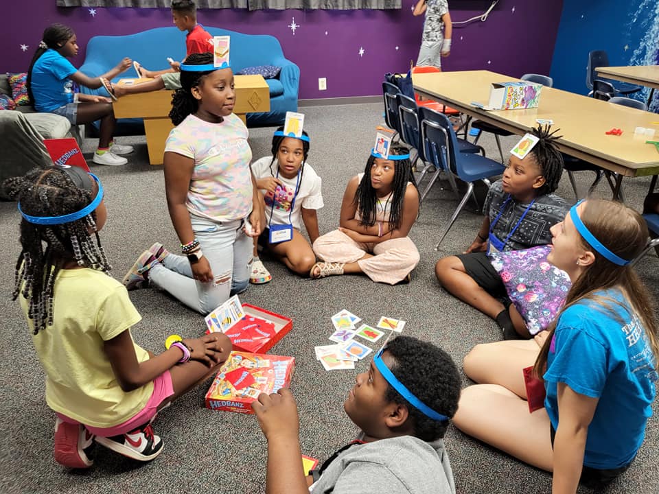 Children playing a game indoors, wearing headbands with cards, gathered around a table.