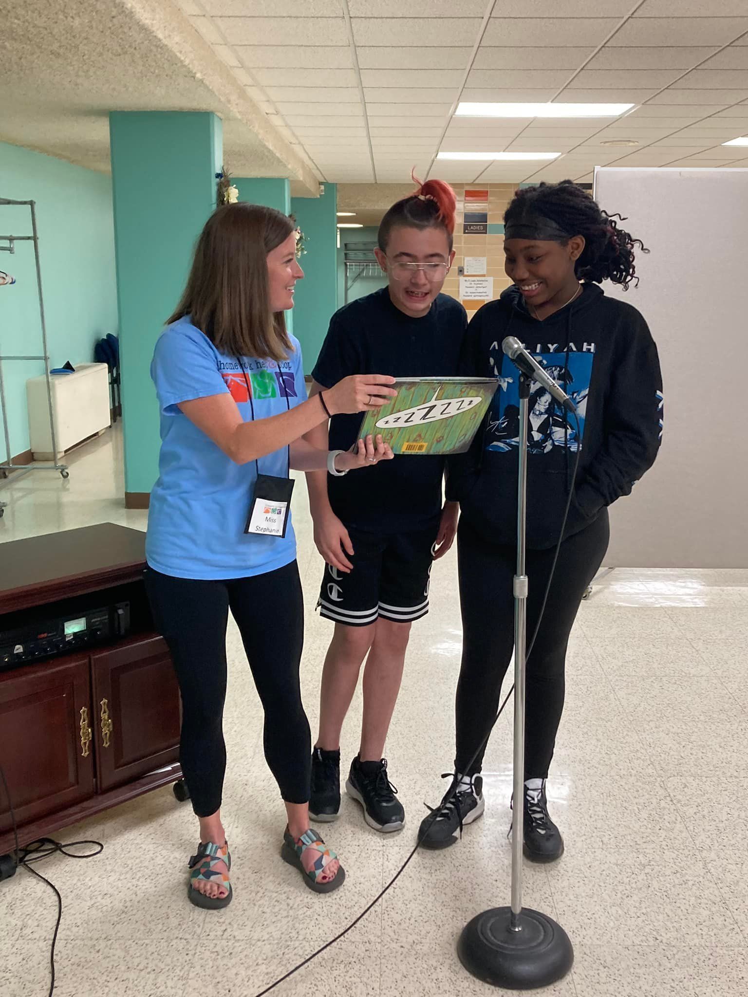 Three people near a microphone: a woman in blue pointing at a book, and two teens, one holding the book, smiling.