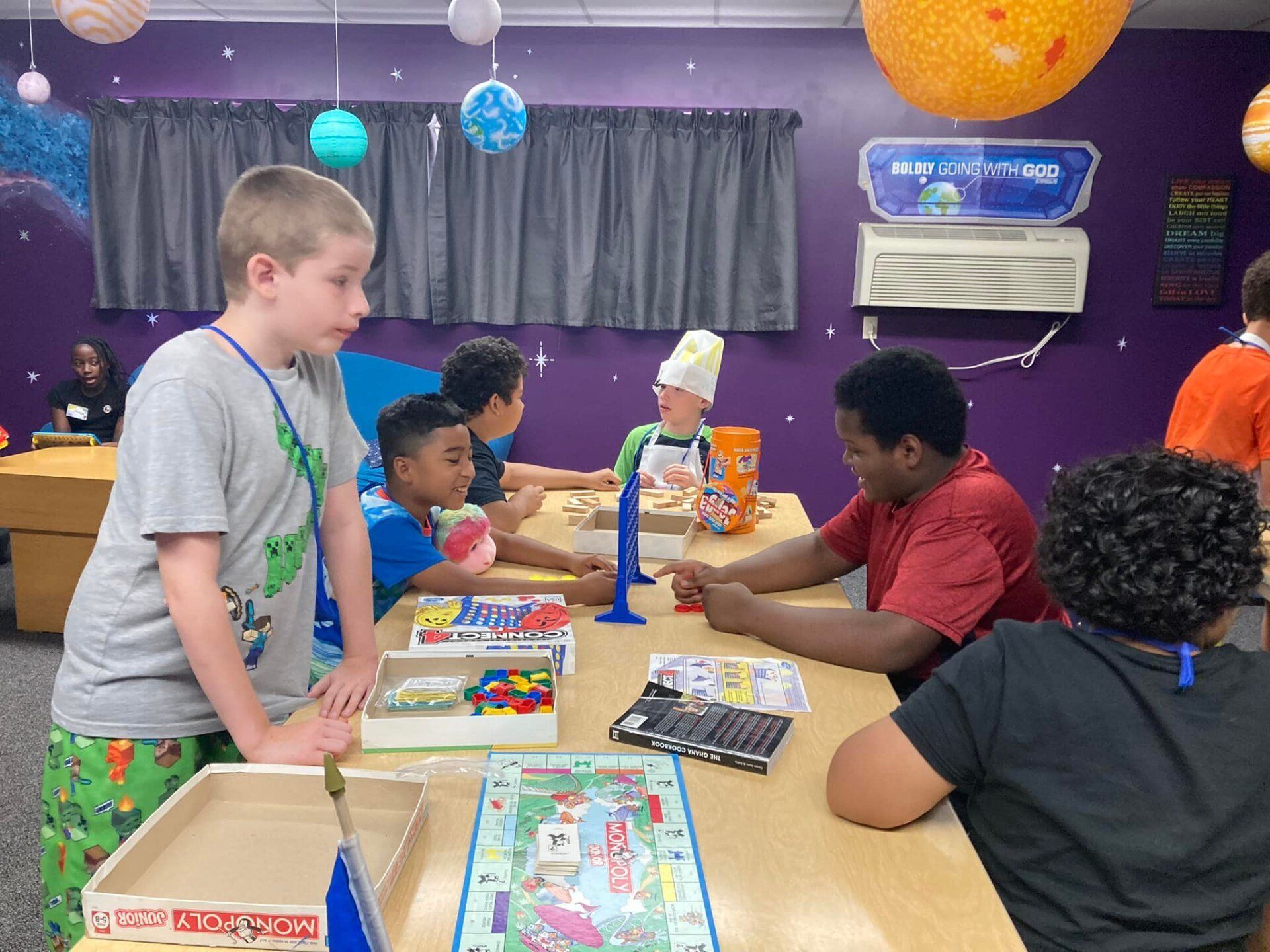 Children playing board games at a table in a colorful room decorated with space-themed elements.