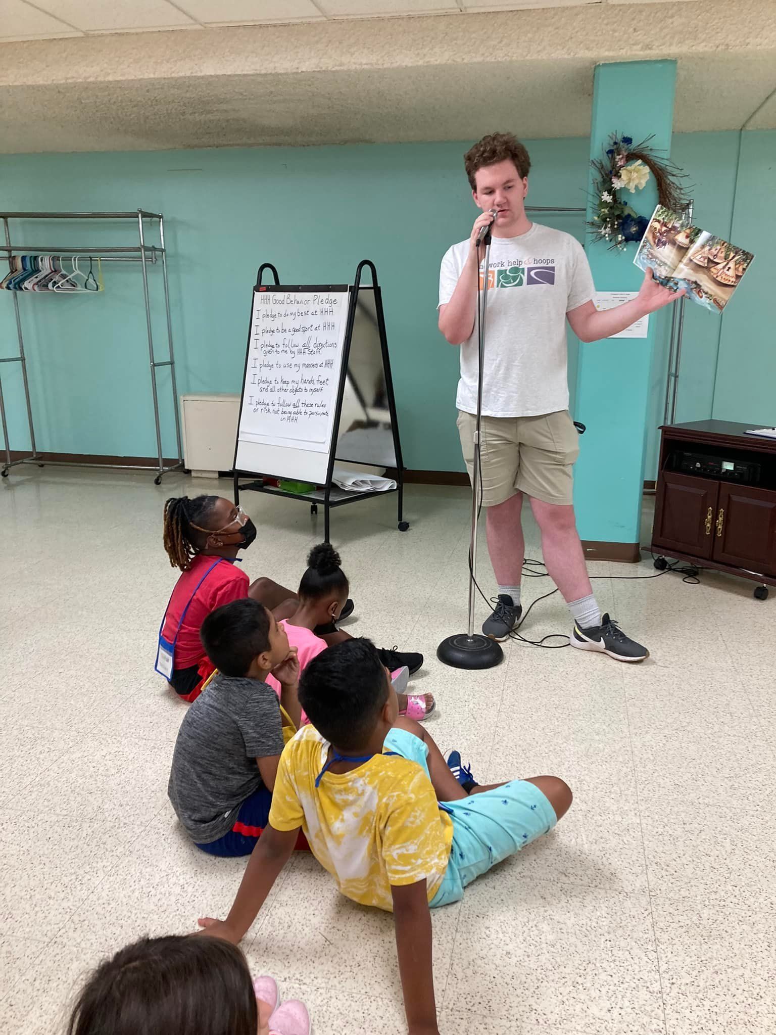 A young man reads a book to a group of children, inside a room with light blue walls.