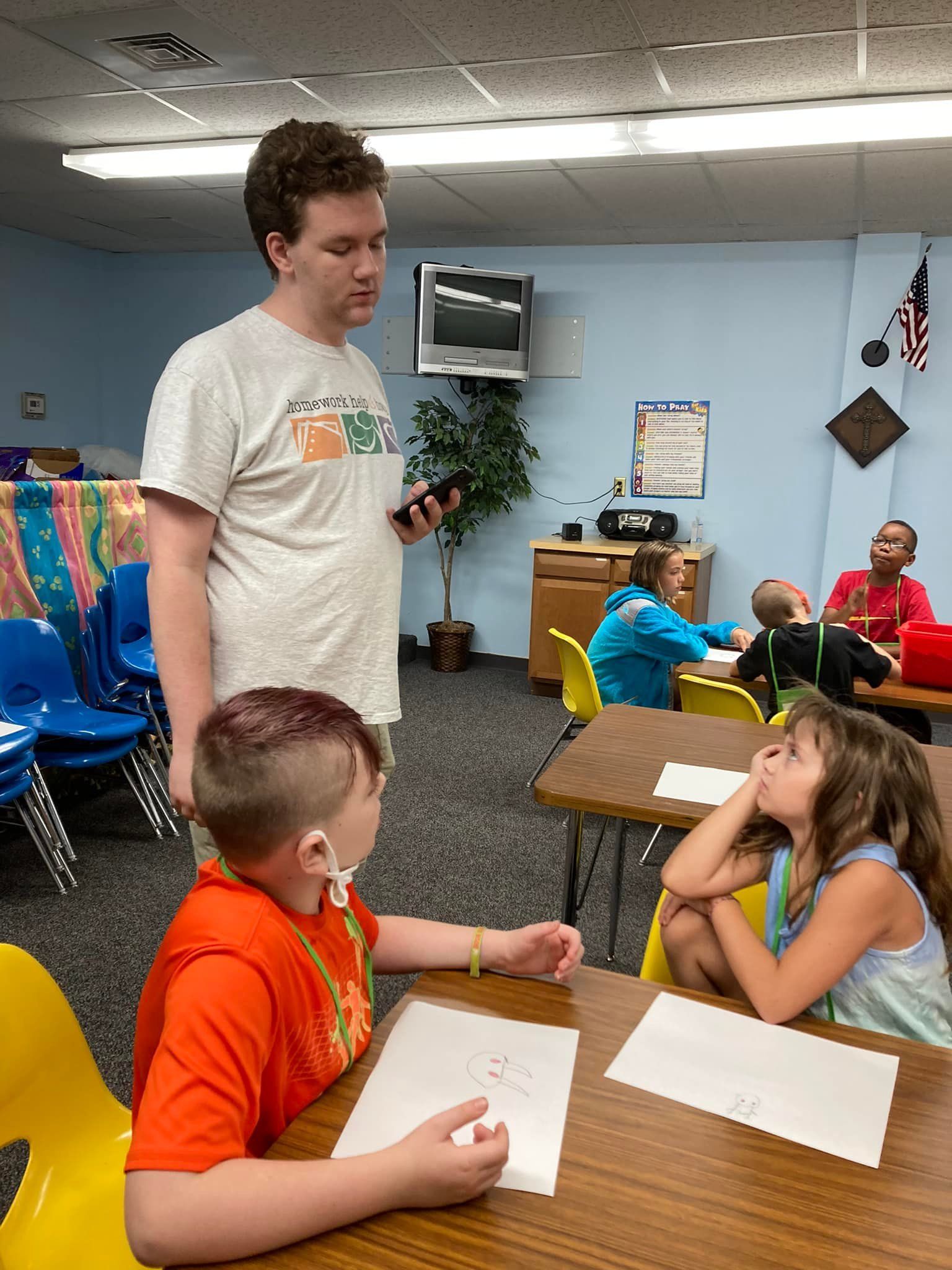 A teacher holds a phone, talking to two students at desks in a classroom. Other kids work nearby.