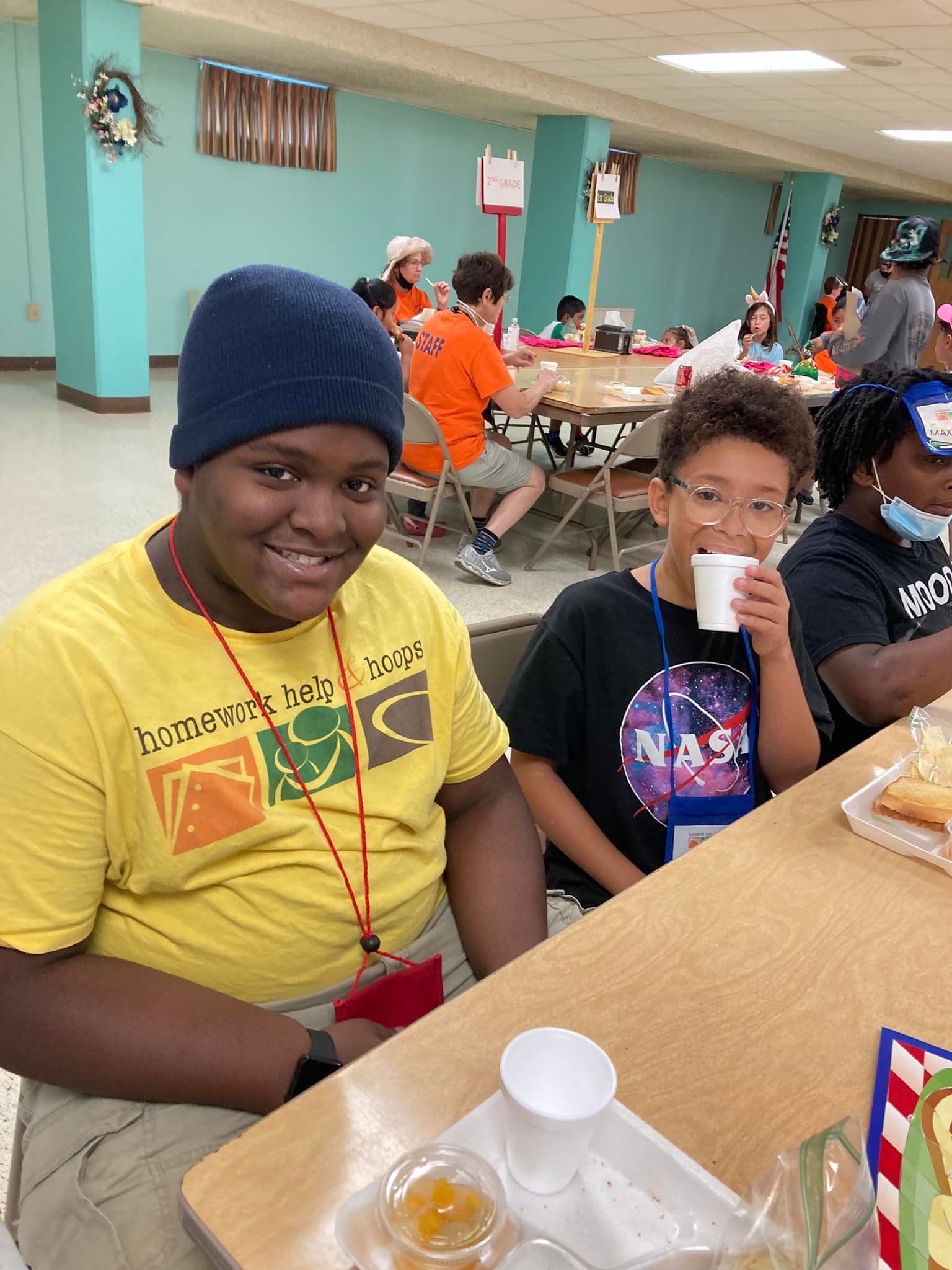 Three kids at a table; one in a blue hat, one in NASA shirt drinking from a cup, and one in a black shirt.