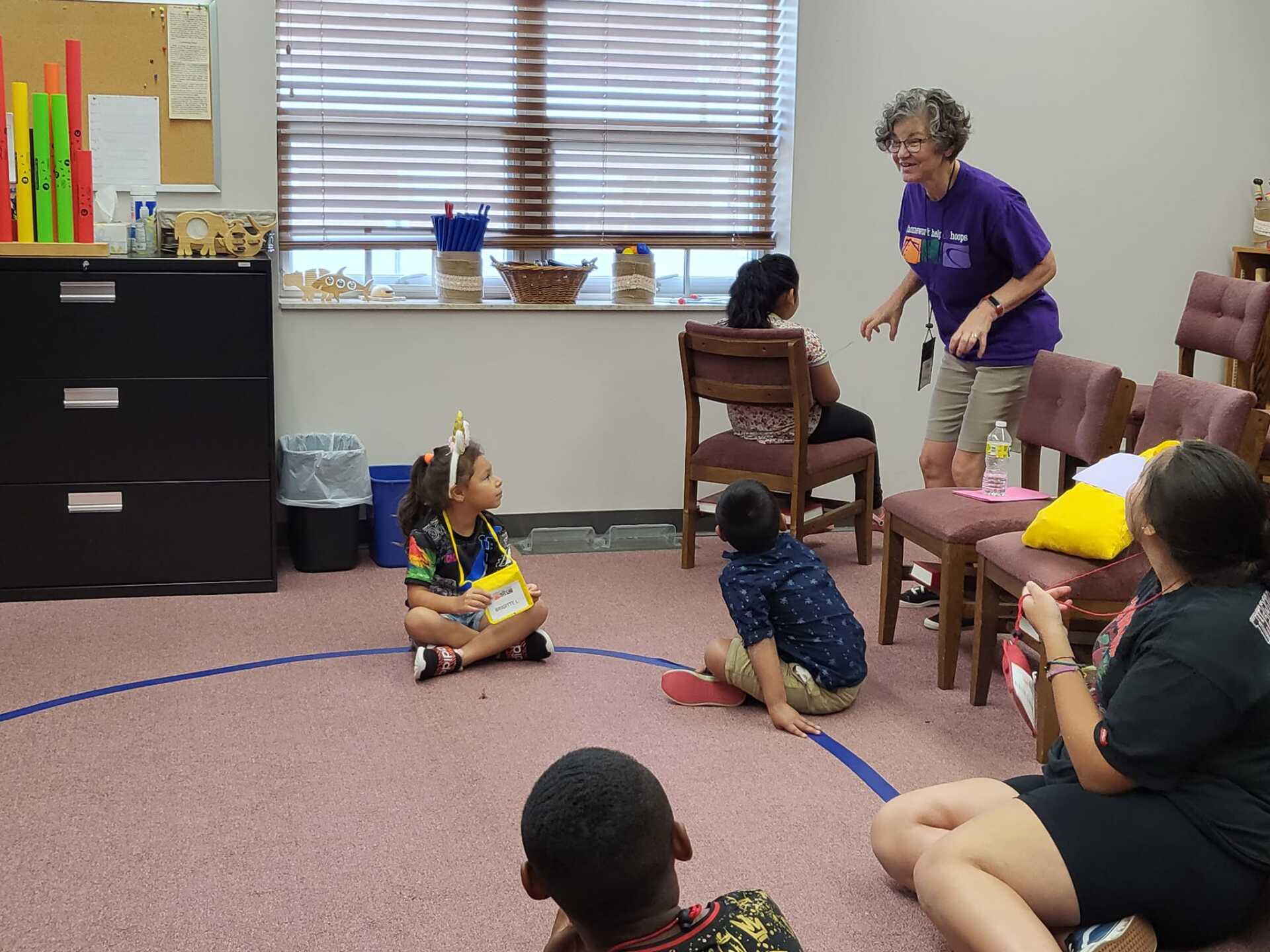 Children and teacher in a classroom with a birthday child, a blue circle on the floor, and a bookshelf.