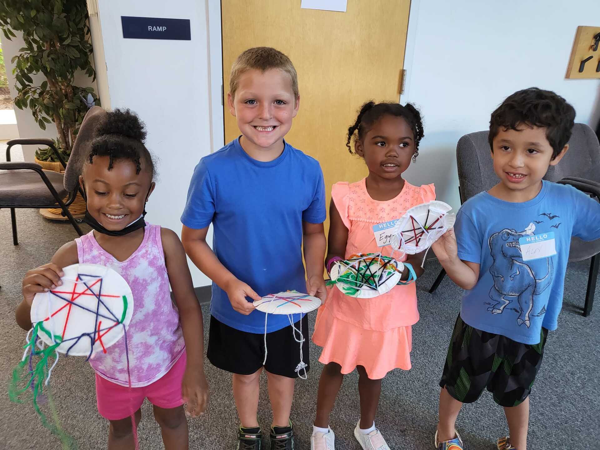 Four smiling children holding colorful crafts, likely in a classroom setting.