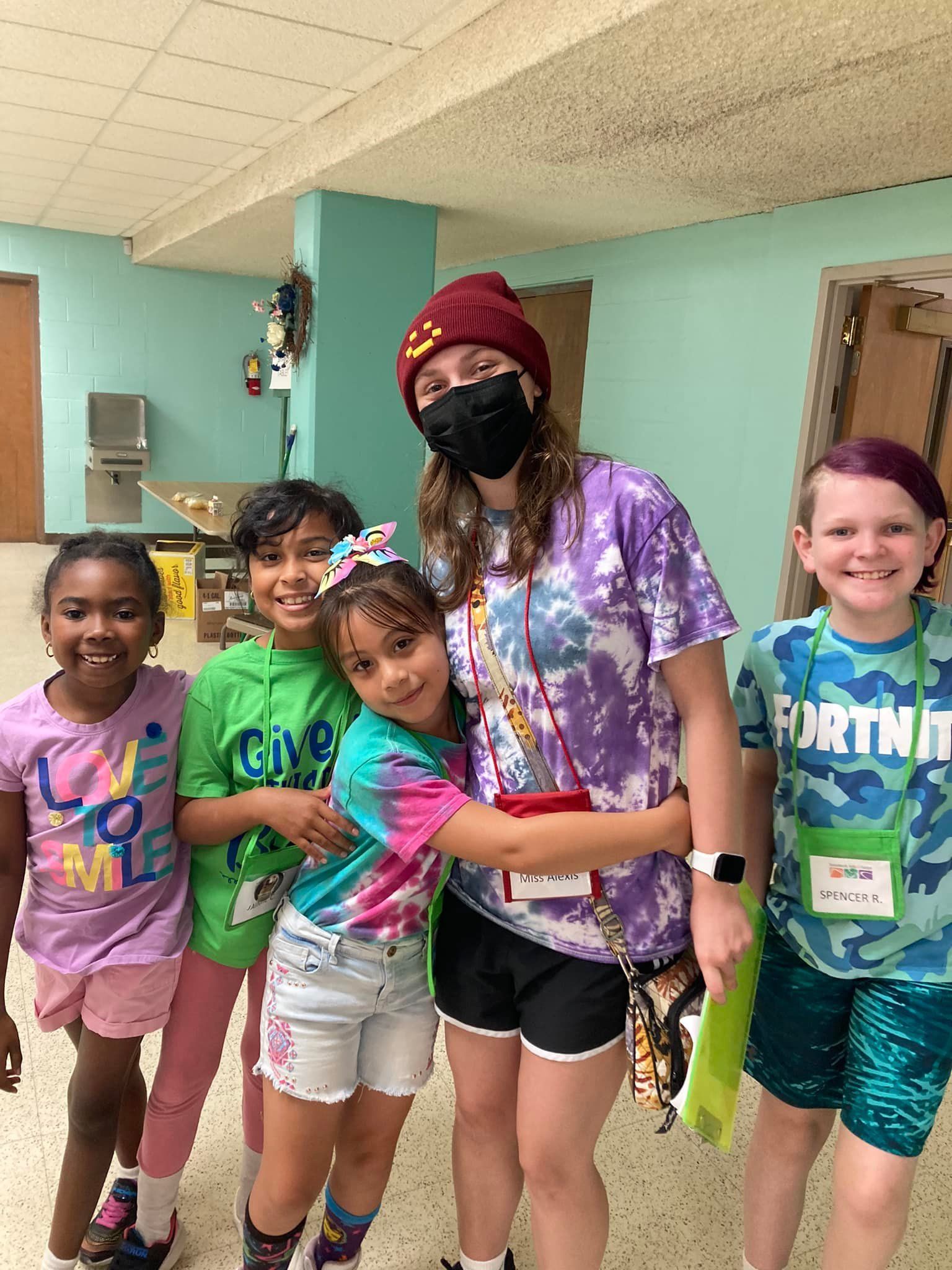 Five children and adult hugging, smiling indoors. Purple tie-dye shirt, beanie, mask.