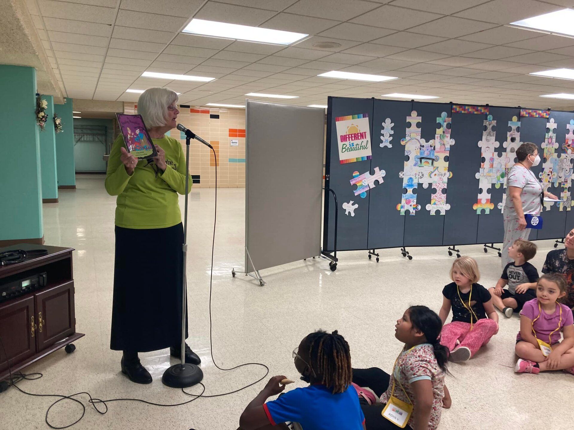 Woman reading to children, holding a book. Setting: school hallway with student art, mic, and sound equipment.