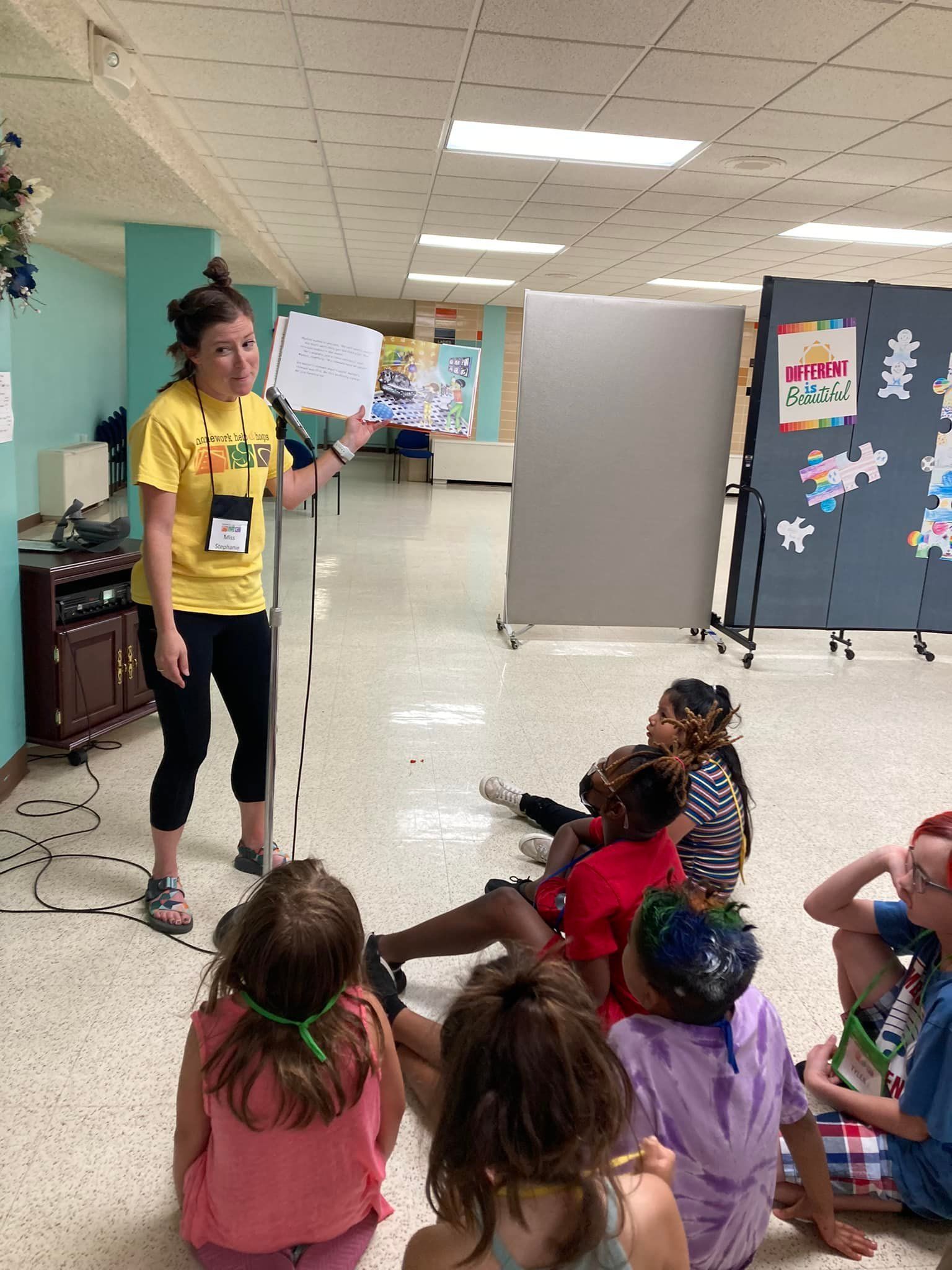 A woman in yellow shirt reads to a circle of children on the floor in a hallway.