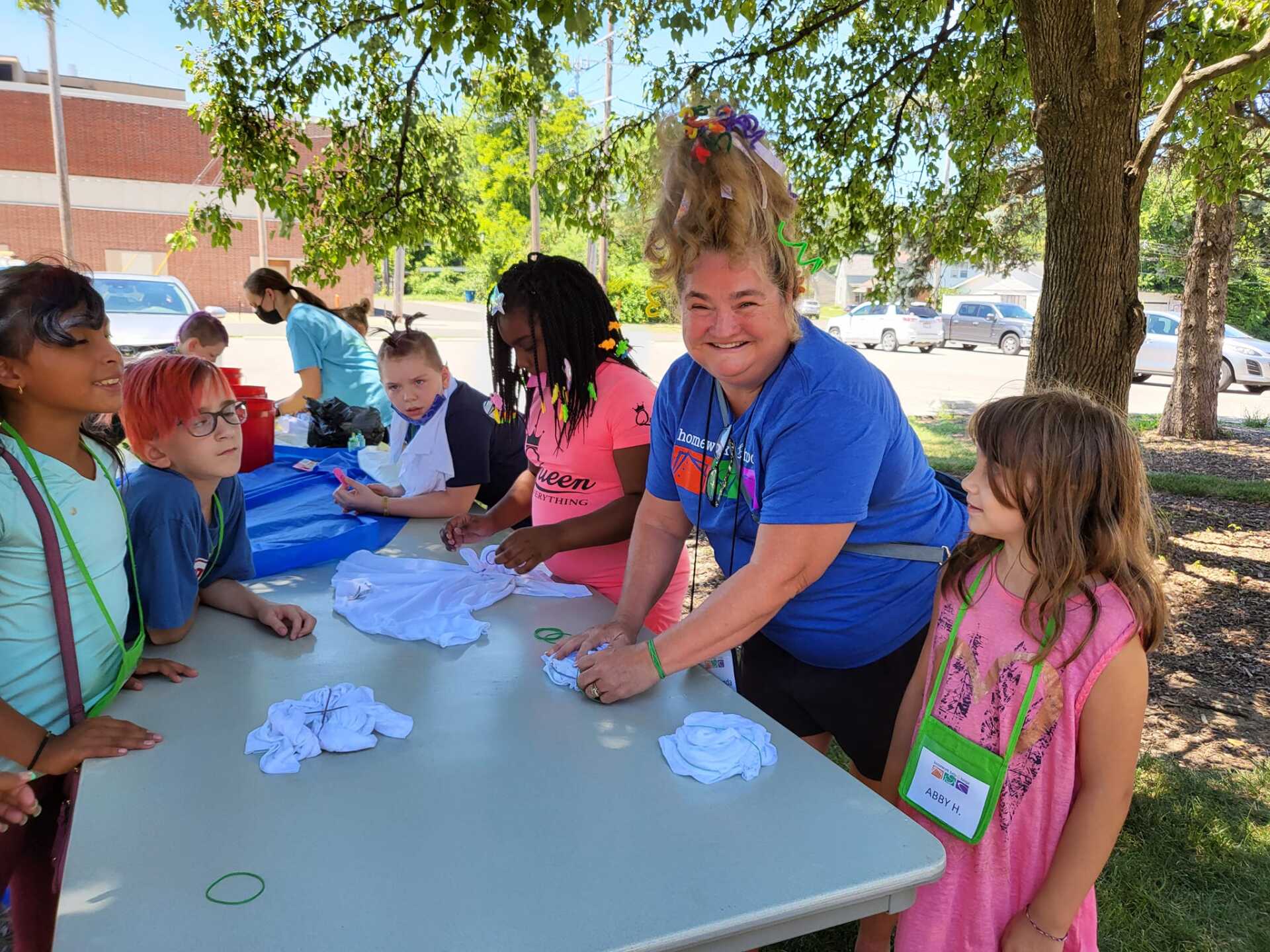 Children and an adult tie-dye shirts at an outdoor table. Kids are gathered around, assisting.