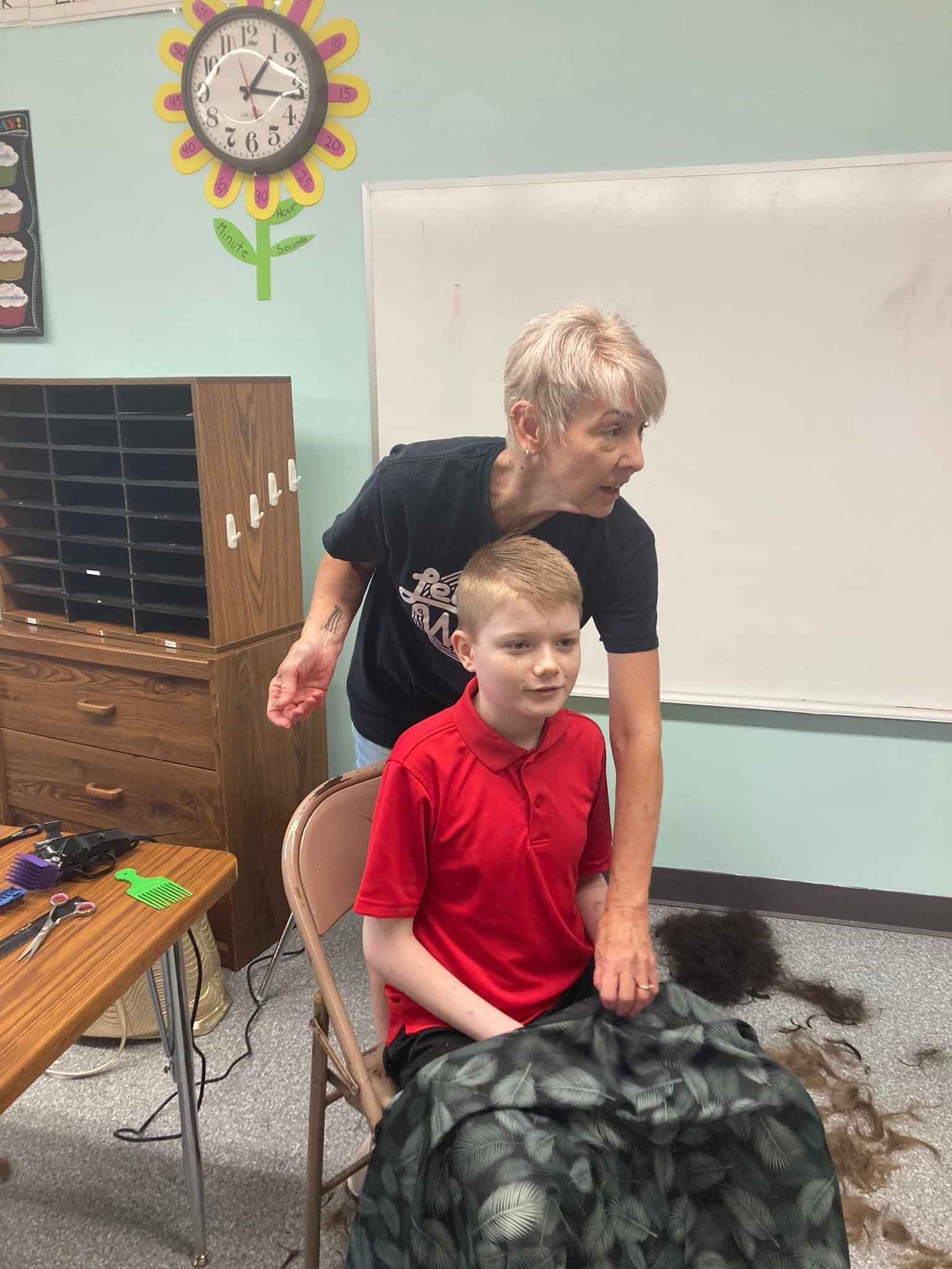 Woman gives a boy a haircut in a classroom; boy is seated, woman is standing.