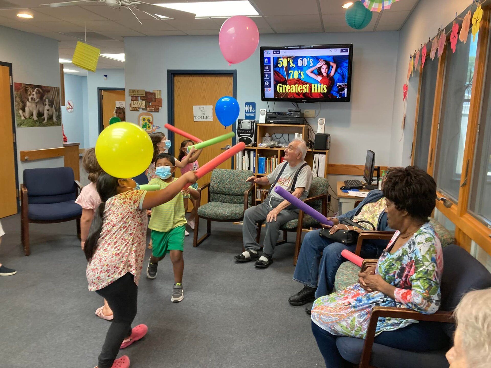 Children and seniors play with foam noodles and balloons in a colorful room, watching a TV.