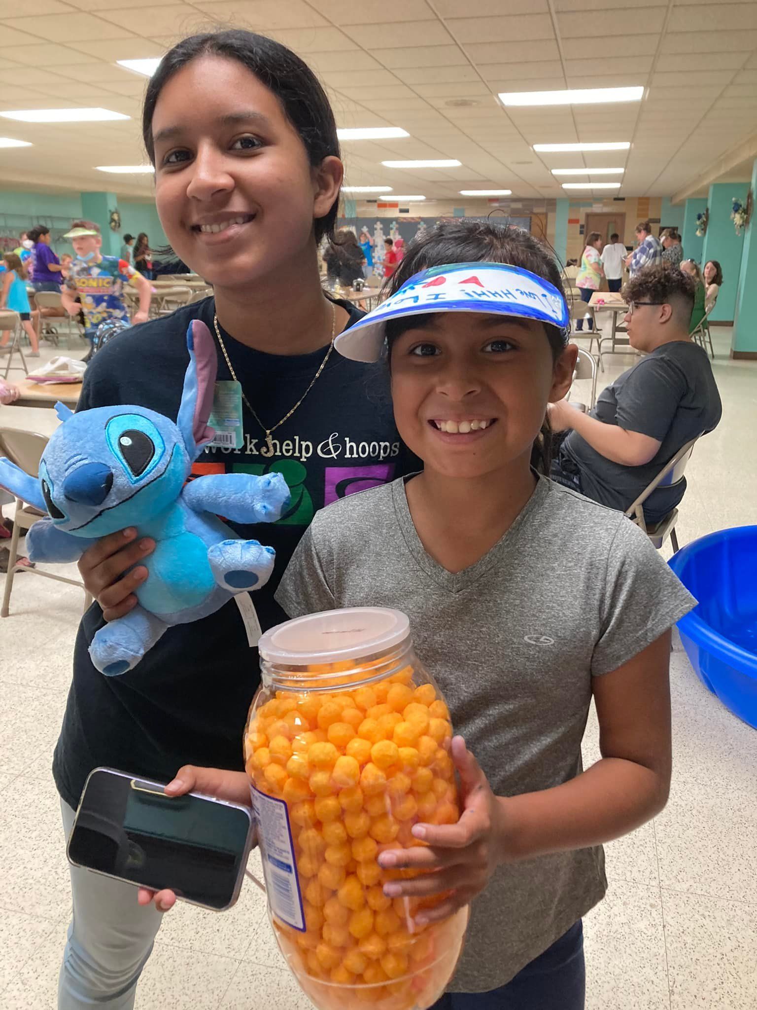 Two smiling girls hold prizes in a school cafeteria; one a stuffed Stitch, the other a jar of cheese balls.