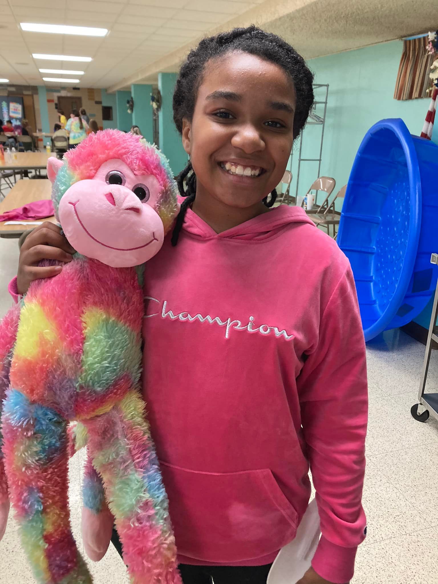 Girl in pink Champion hoodie smiles, holding a colorful stuffed monkey. Indoor setting.