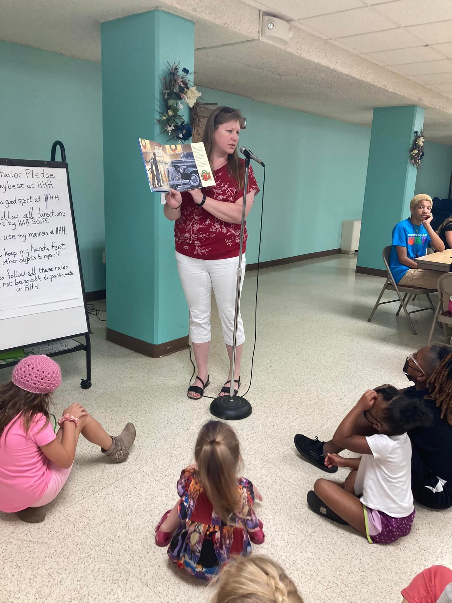 Woman reading a book to children. She stands by a microphone. Children sit on the floor. Light blue wall.